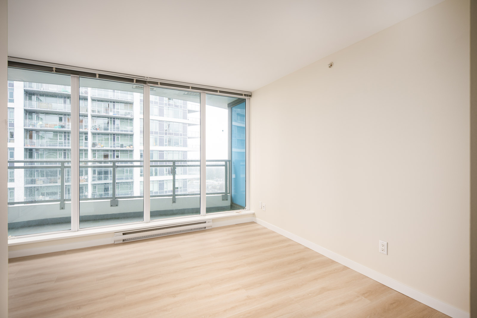 Unfurnished room with light wood flooring, large floor-to-ceiling windows, and a balcony overlooking modern apartment buildings.