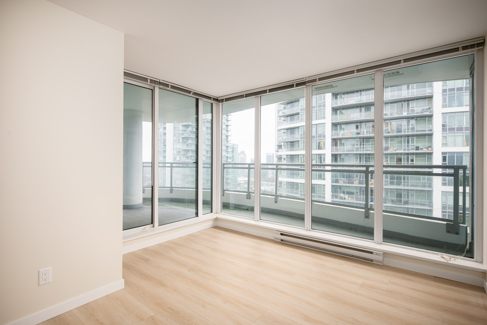 Empty modern apartment room with light wood floors, white walls, and floor-to-ceiling windows overlooking neighboring high-rise buildings and a balcony.