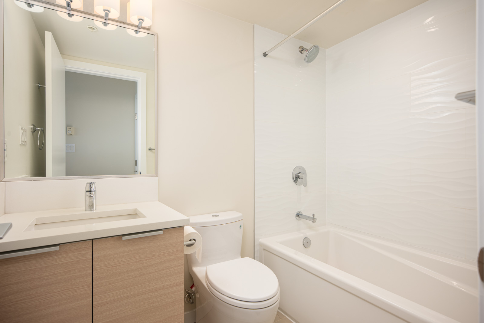 Modern bathroom with light wood vanity, white sink, toilet, and bathtub-shower combination with white tile. Large mirror and overhead lighting above the sink.
