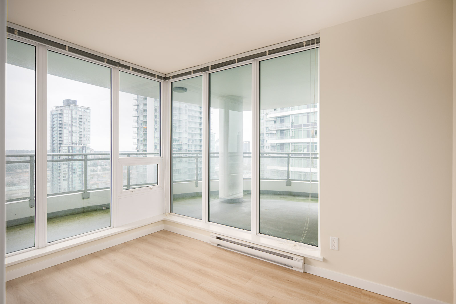 Empty corner room with light wood flooring, floor-to-ceiling windows, and a view of high-rise buildings and a covered balcony outside.