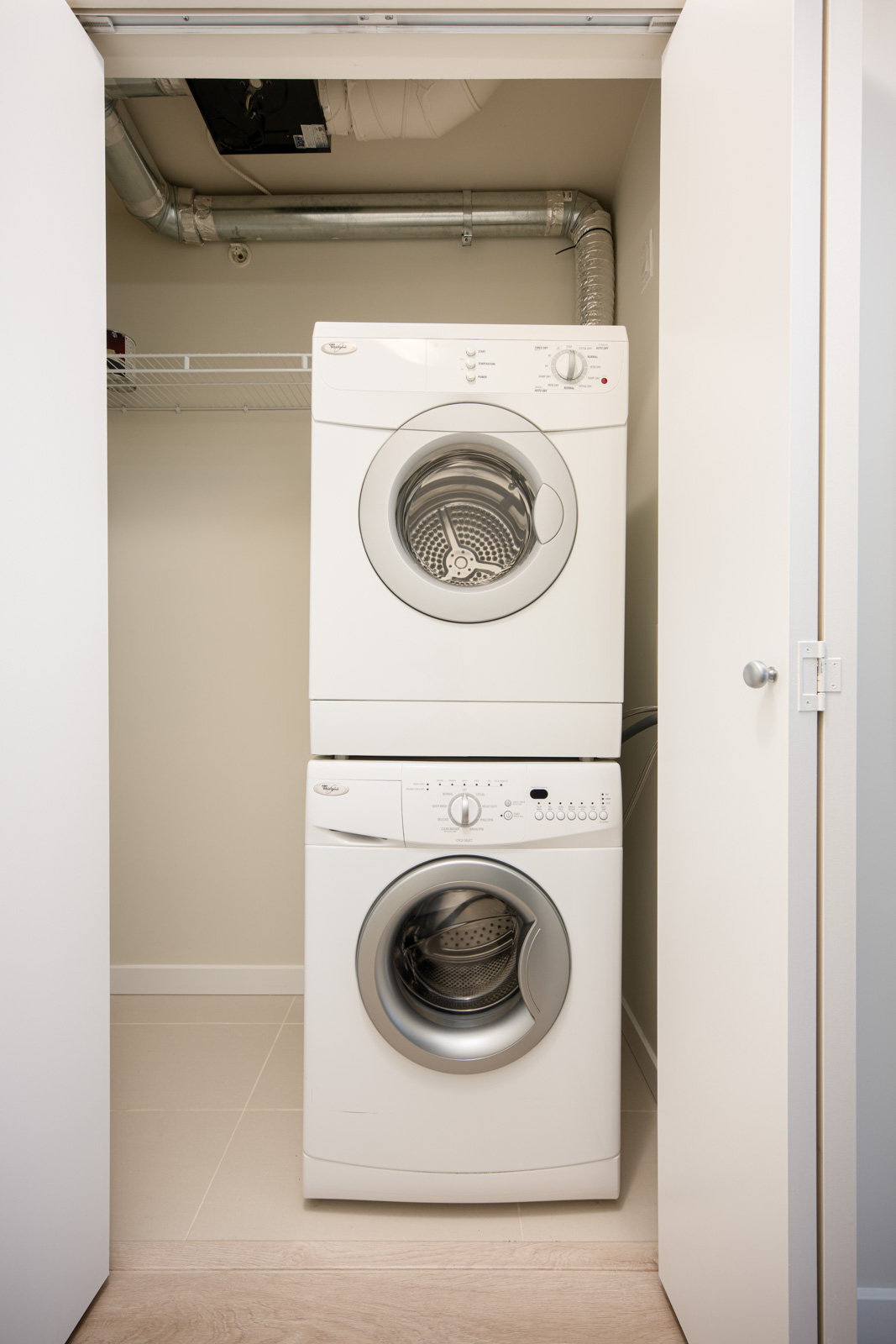 Stacked front-loading washer and dryer units in a small closet with open doors, exposed vent pipes, and a wire shelf above the machines.