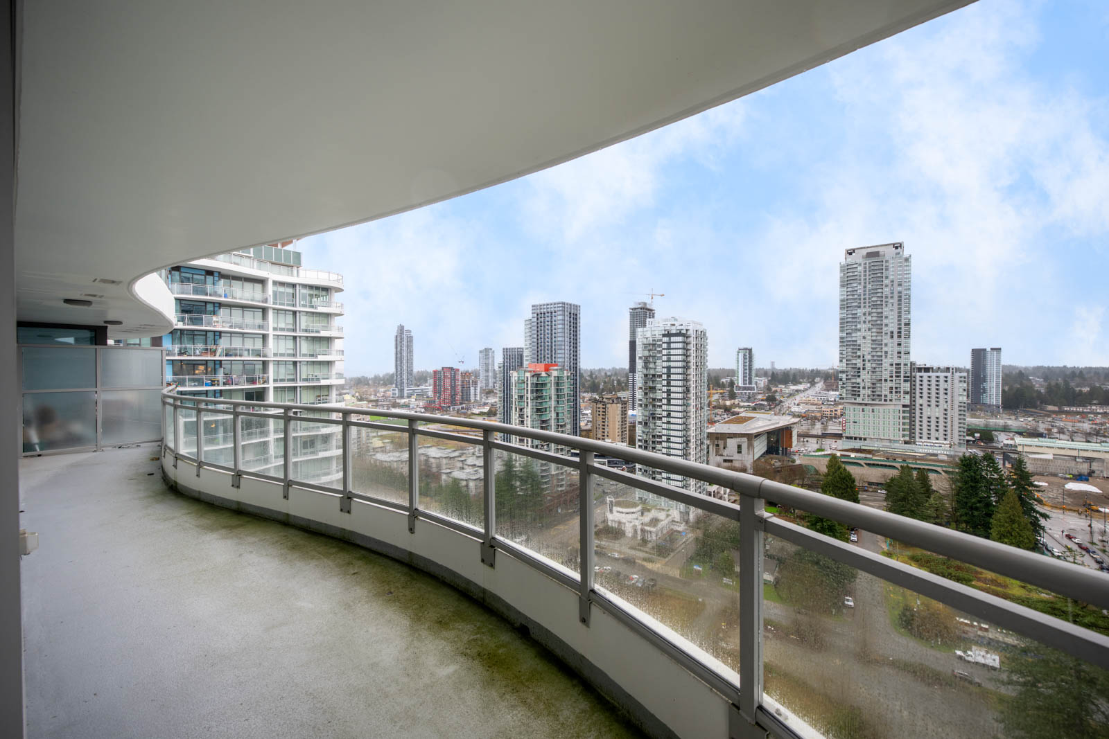 Curved high-rise balcony with glass railing overlooking a cityscape of tall buildings and a cloudy sky.