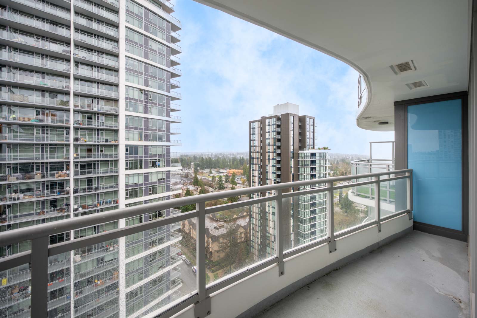 View from a high-rise balcony showing neighboring modern apartment buildings with glass facades and a partly cloudy sky in the background.