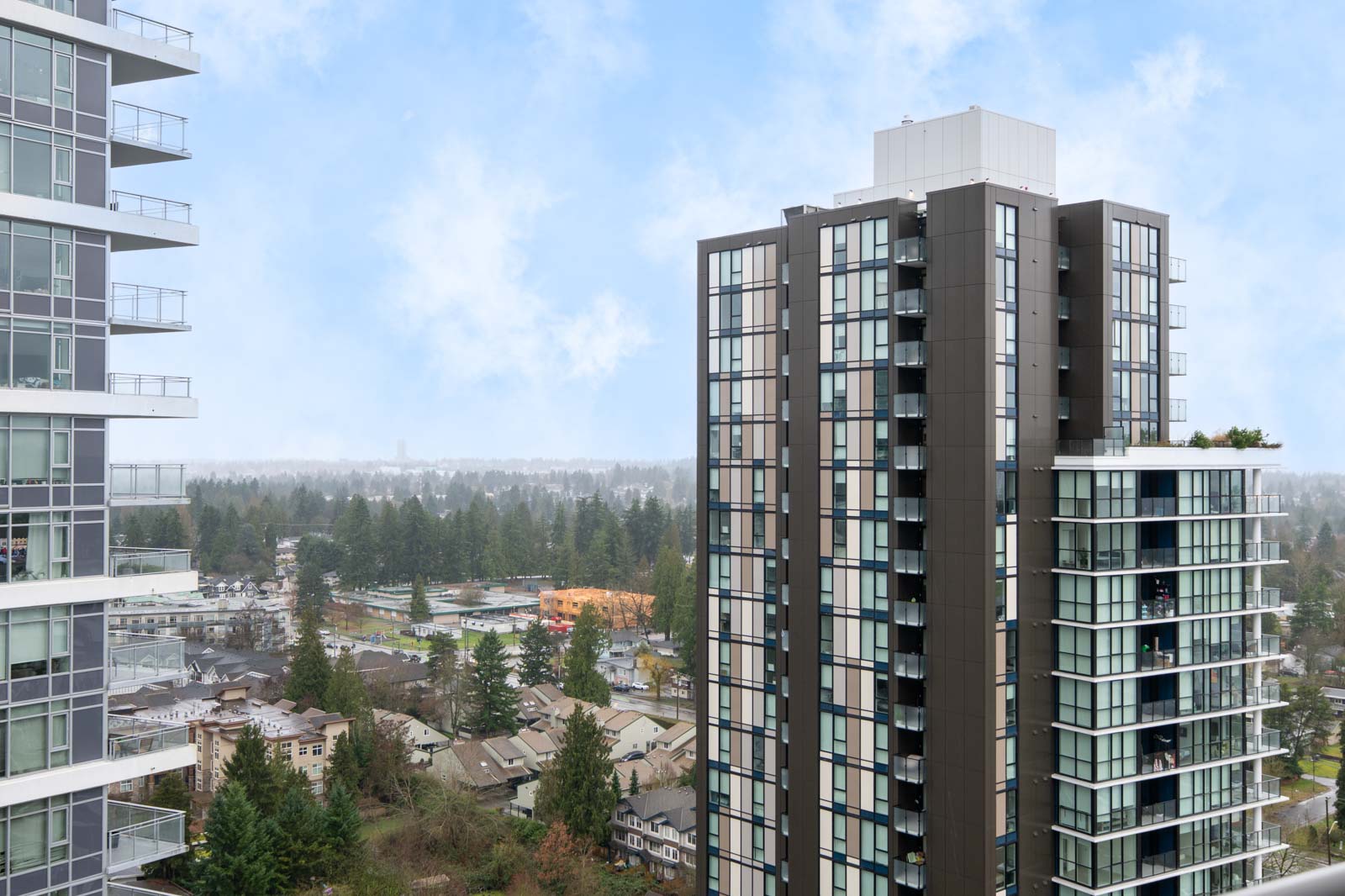 Two modern high-rise apartment buildings with glass balconies overlook a residential neighborhood and trees under a partly cloudy sky.