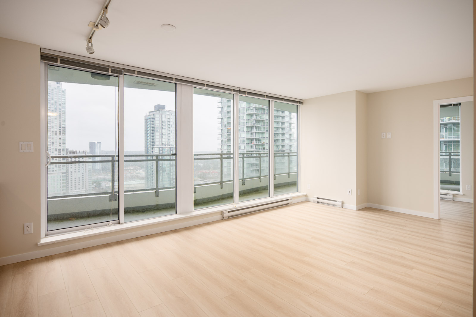 Unfurnished modern apartment with light wood flooring, large floor-to-ceiling windows, and a balcony overlooking city buildings on a cloudy day.