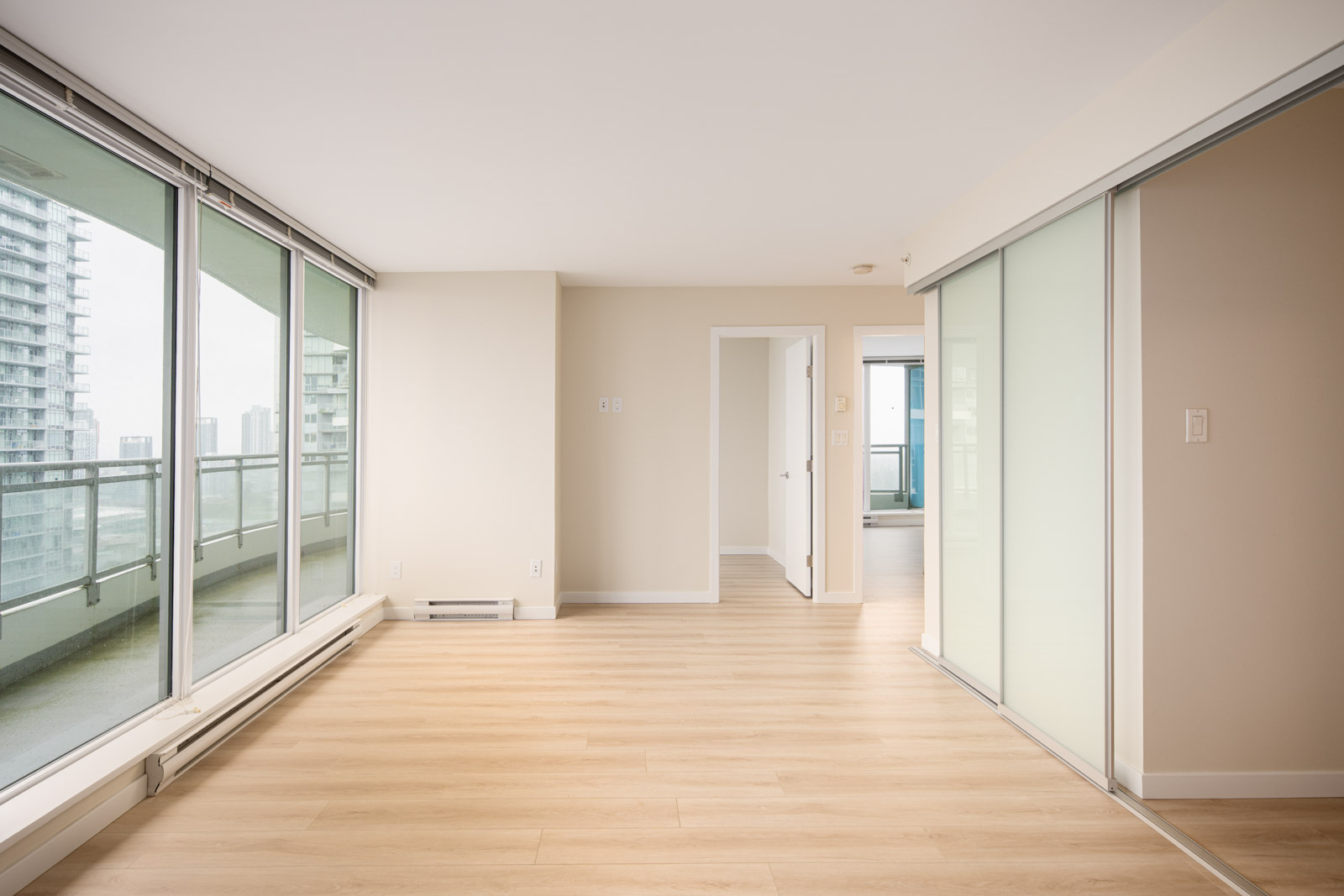 Bright, empty apartment room with light wood floors, large floor-to-ceiling windows, glass sliding doors, and a view of city buildings outside.