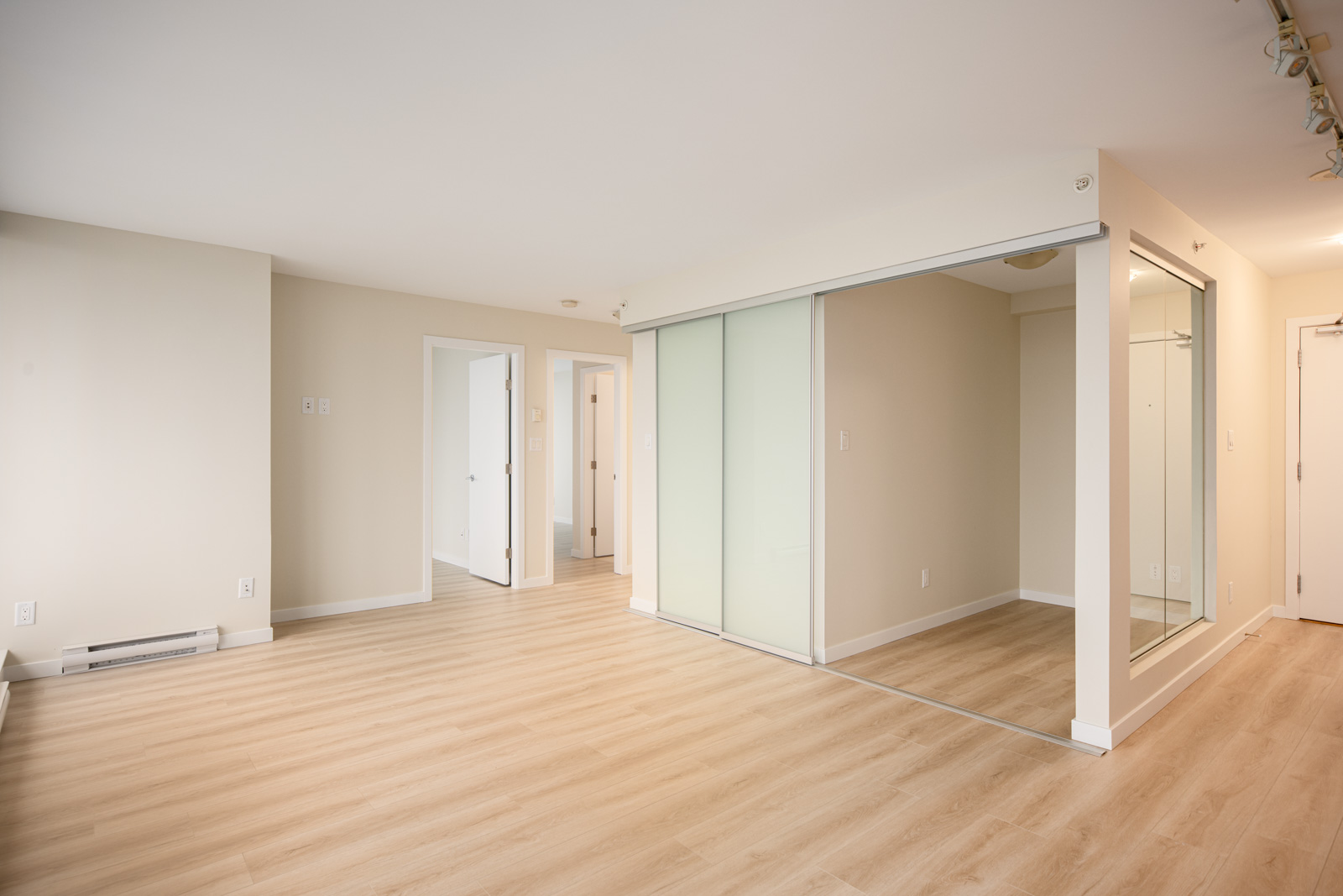 A bright, empty room with light wood flooring, beige walls, and a glass partition separating a small area; two white doors lead to adjacent rooms.