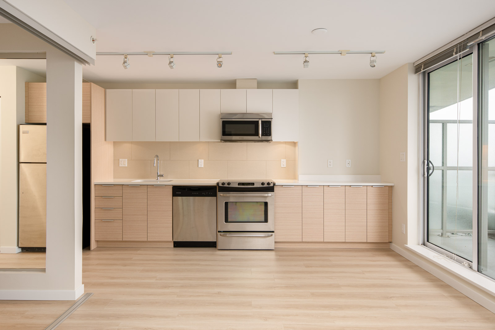 Modern kitchen with light wood cabinets, stainless steel appliances, white countertops, and large windows providing natural light.
