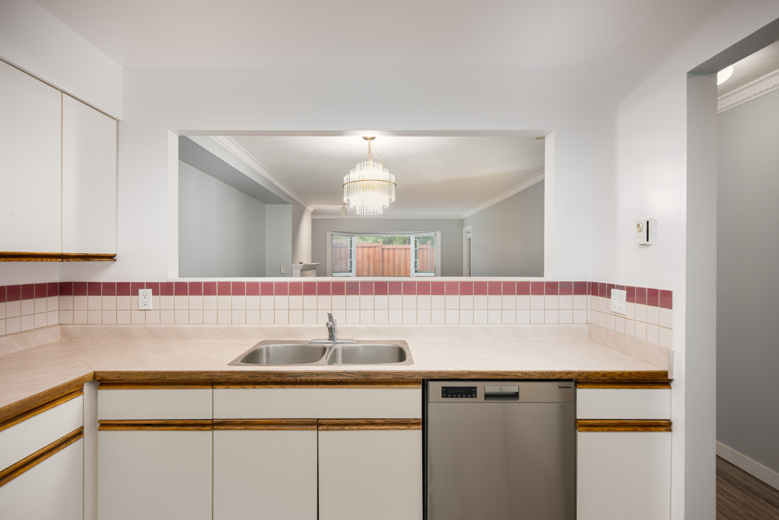 A kitchen with white cabinets, a double sink, a dishwasher, and a pass-through window overlooking a dining area with a chandelier.