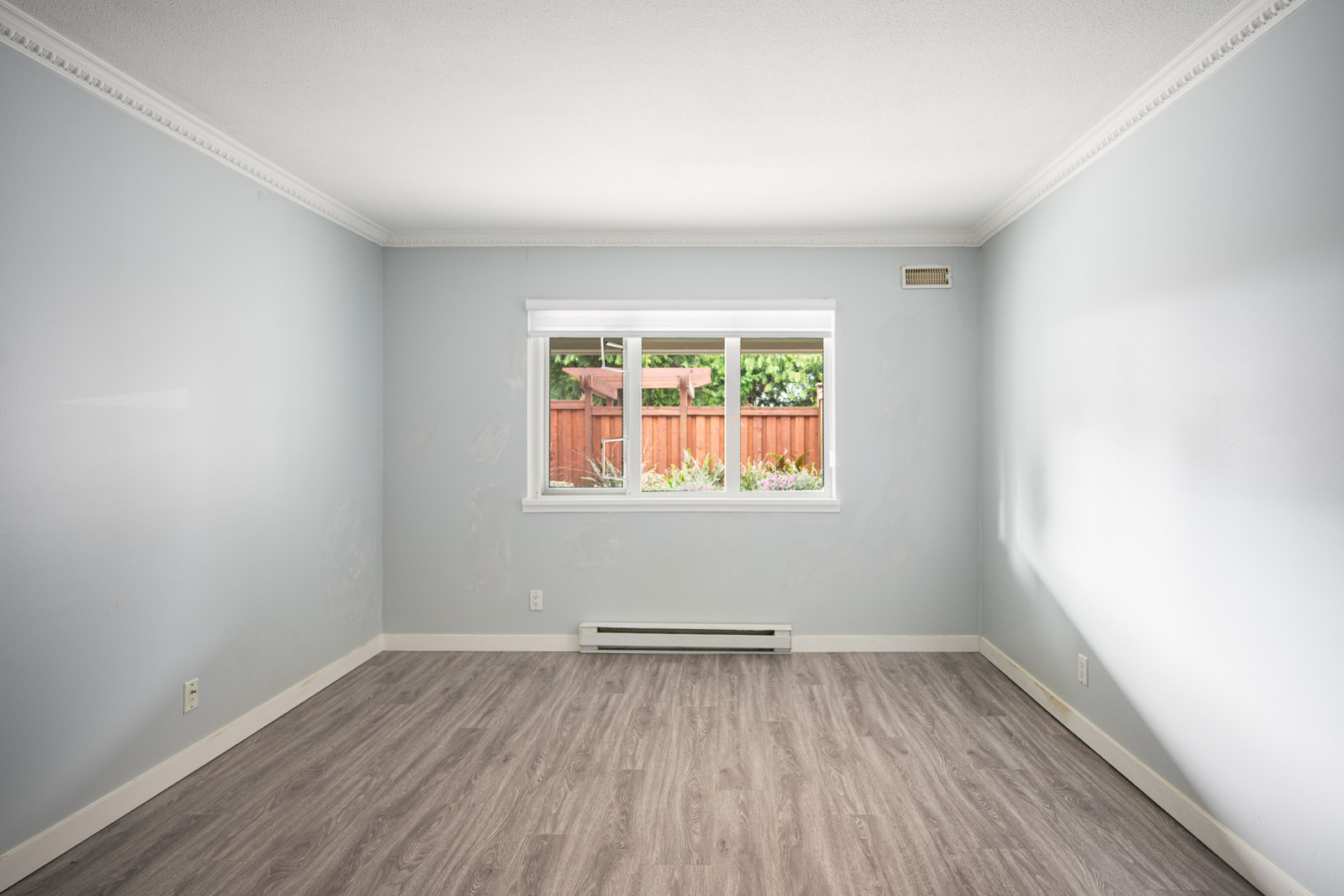 Empty room with light gray walls, white trim, and wood-style flooring, featuring a large window letting in natural light and a view of a wooden fence and greenery outside.