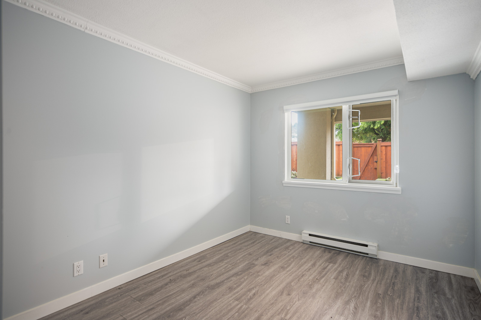 Empty room with light blue walls, white trim and baseboard heater, wood-style flooring, and a window showing an outdoor area with a fence and greenery.