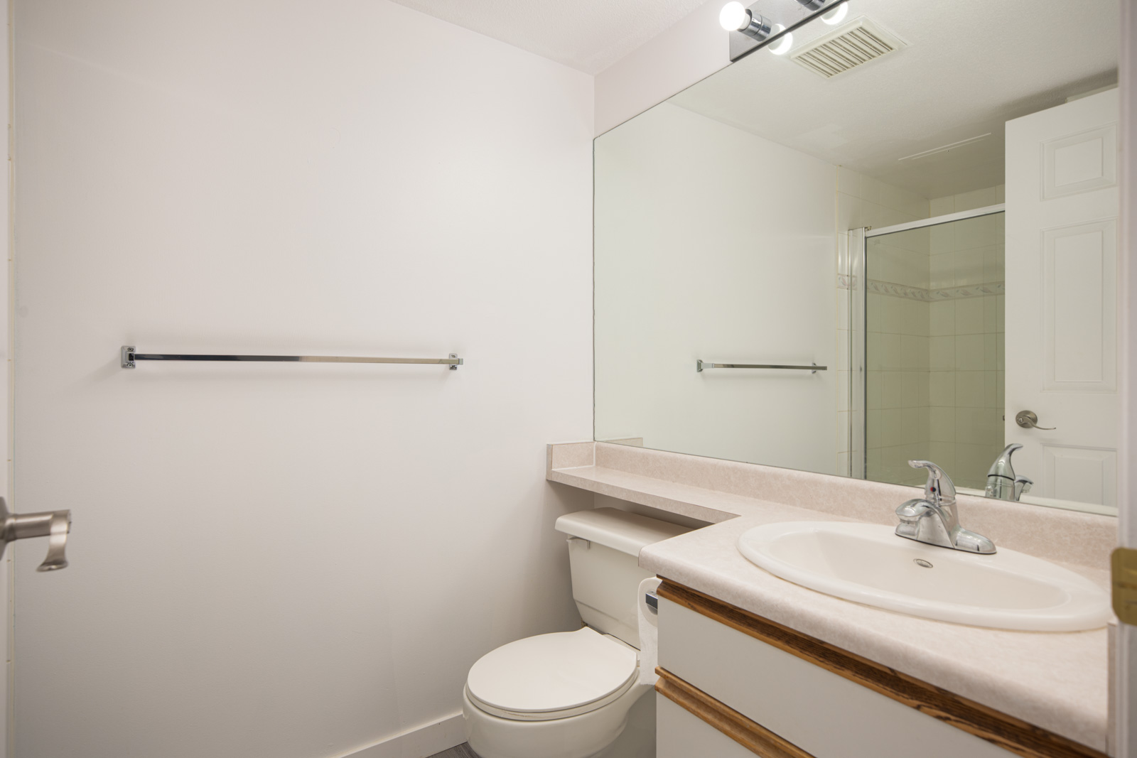 A simple bathroom with a sink, countertop, toilet, large wall mirror, towel rack, and a glass shower enclosure.