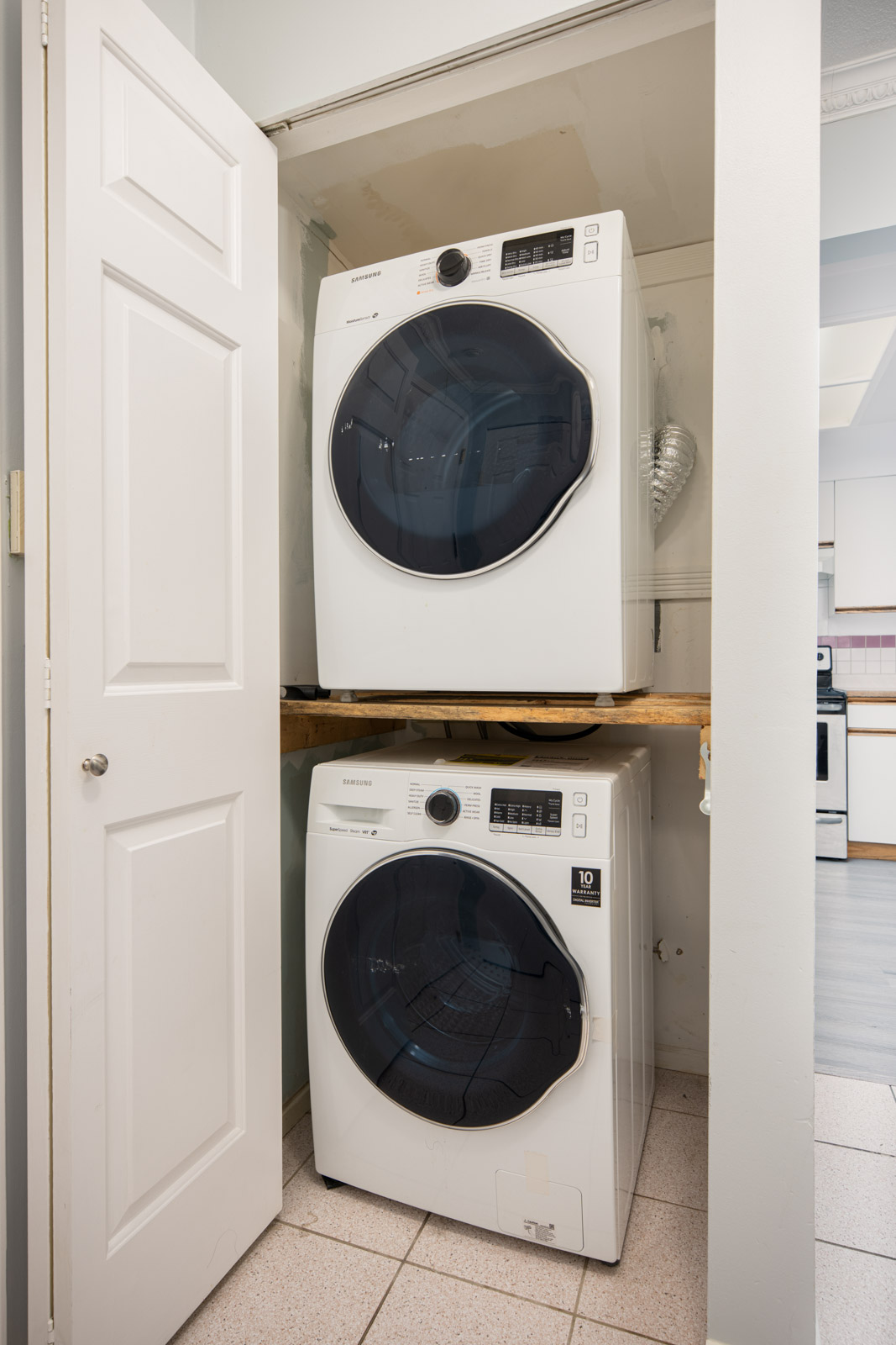 A stacked washer and dryer set inside a small laundry closet with shelves, partially open white door, and vent hose visible behind the units.