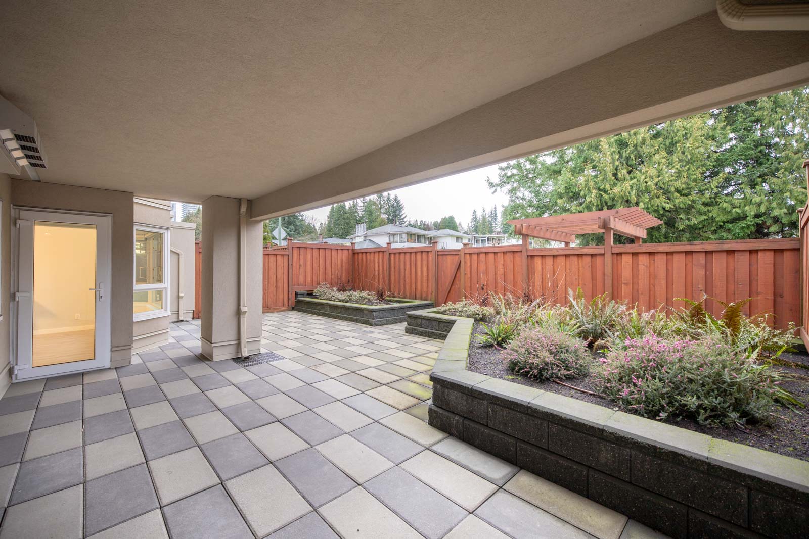 Covered patio with tiled flooring, raised garden beds, and a wooden fence surrounding the backyard. A glass door leads inside the house.