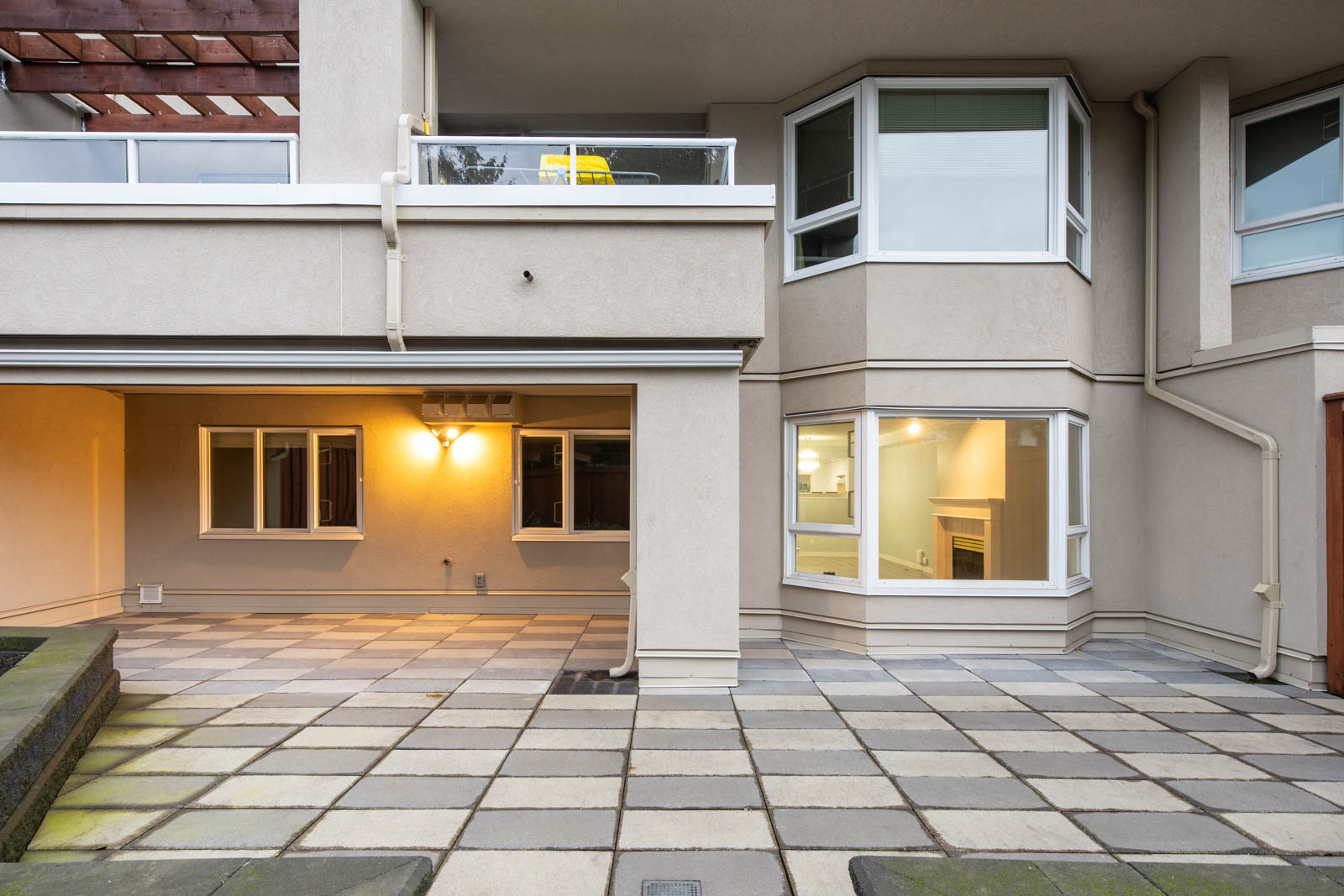 Modern beige apartment exterior with large windows, balcony, covered patio area, and square-tiled courtyard.