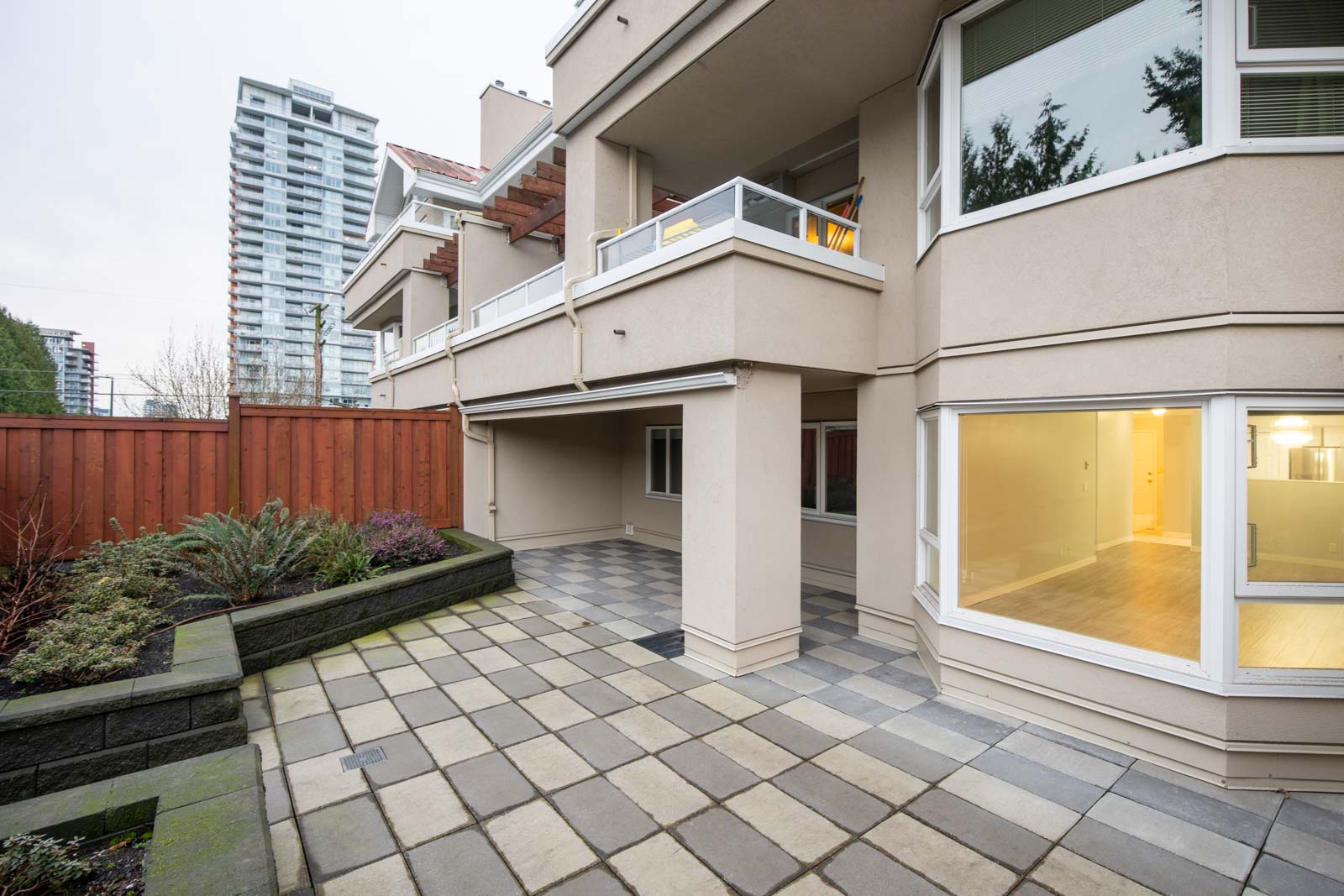 Ground-level patio area with tiled flooring, beige walls, large windows, and balcony above; landscaped garden and wooden fence on the side, with a high-rise building in the background.