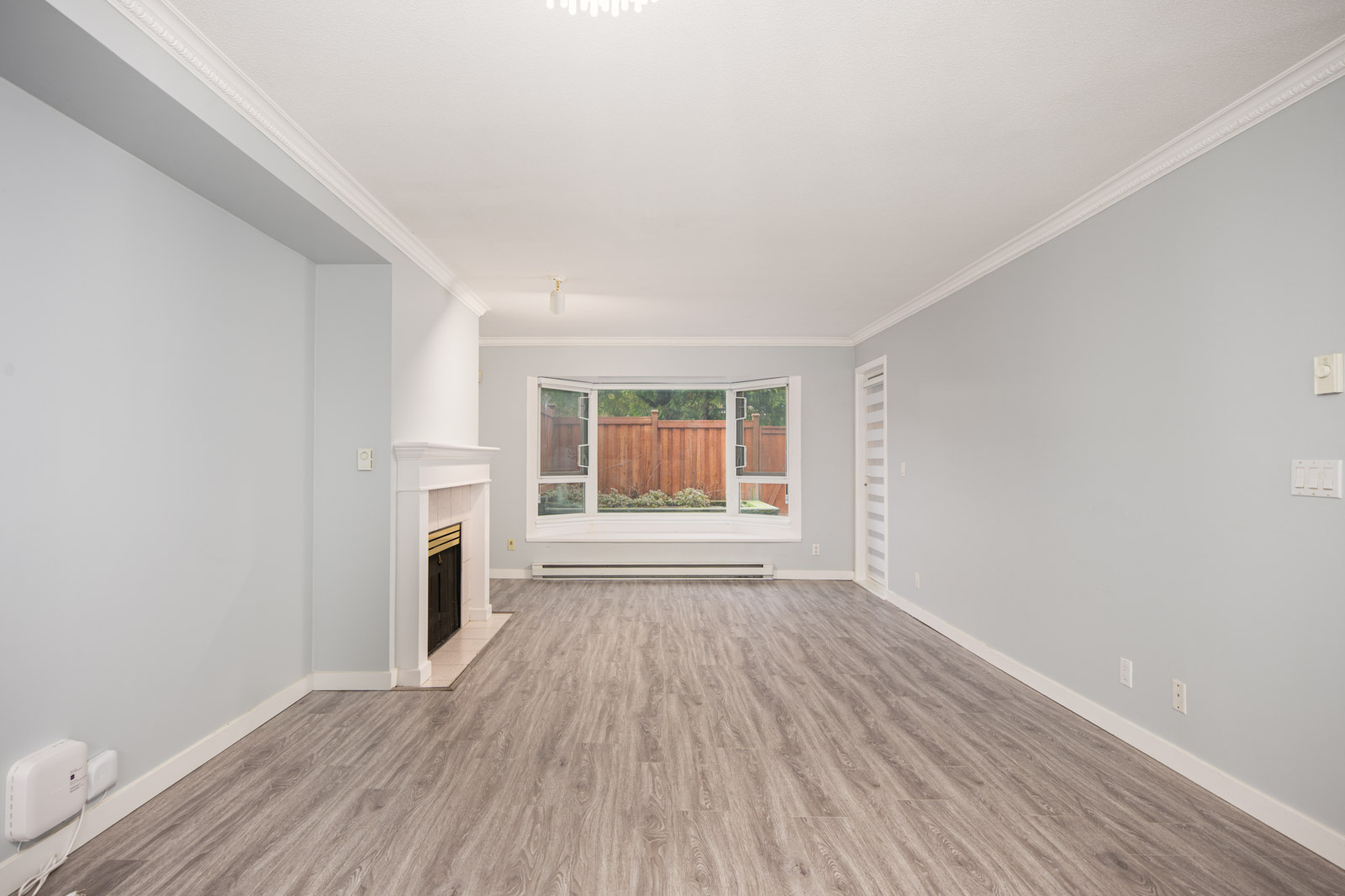 Unfurnished living room with light gray walls, wood-look flooring, a white fireplace, large window, and view of a fenced backyard.