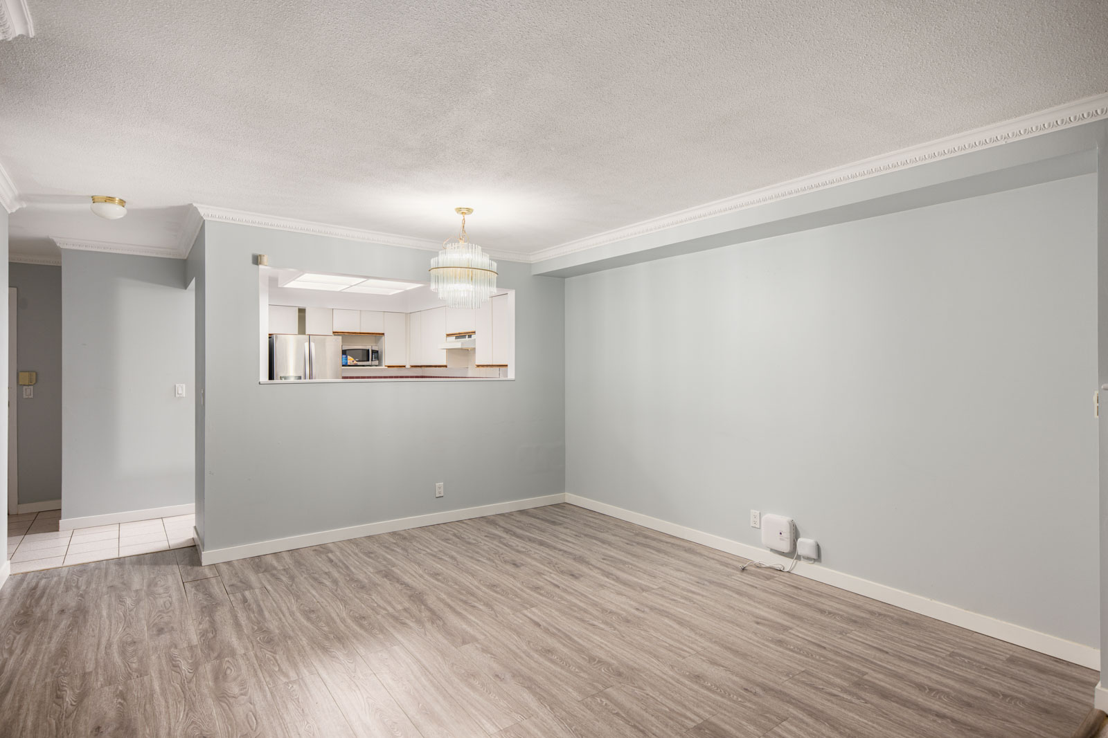 Unfurnished living room with light gray walls, wood flooring, a ceiling light fixture, and a view into the adjacent kitchen with stainless steel appliances.