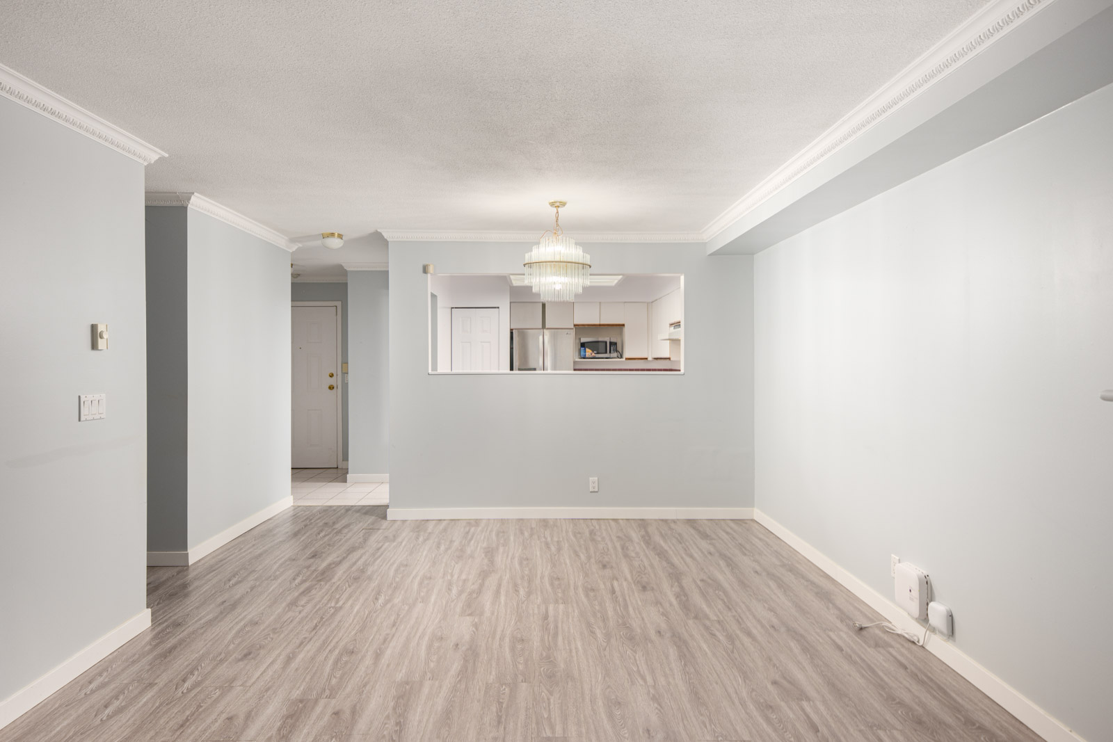 Empty living room with light gray walls, laminate wood flooring, and a view into a kitchen through a pass-through opening. A ceiling light fixture hangs in the center.