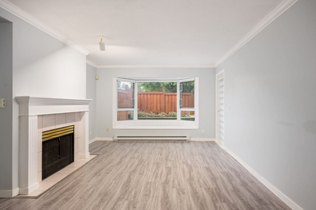 Empty living room with light gray walls, a white fireplace on the left, large bay window facing a fenced yard, and wood-look flooring.