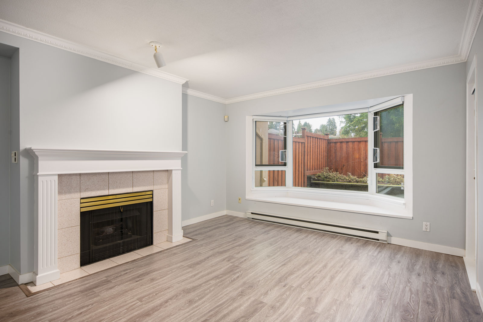 Unfurnished living room with a fireplace, large bay window, light grey walls, and wood-look flooring. A wooden fence and greenery are visible outside the window.