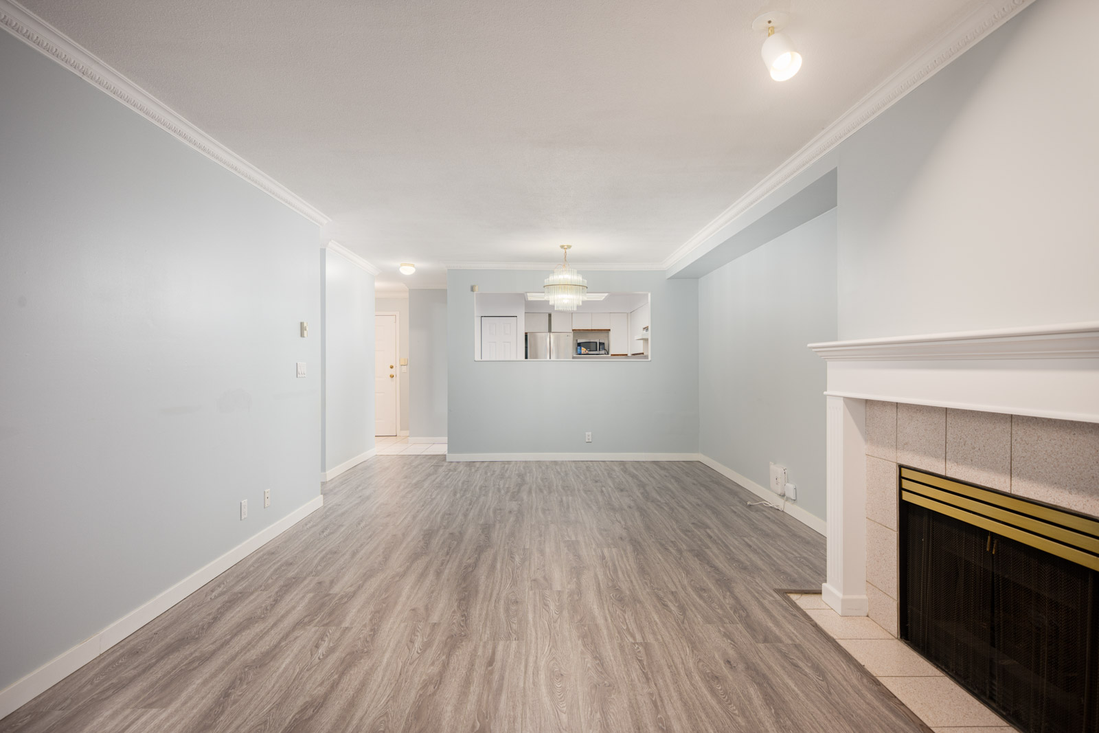 Unfurnished living room with light gray walls, wood-look flooring, a fireplace on the right, and an open pass-through to the kitchen in the back.