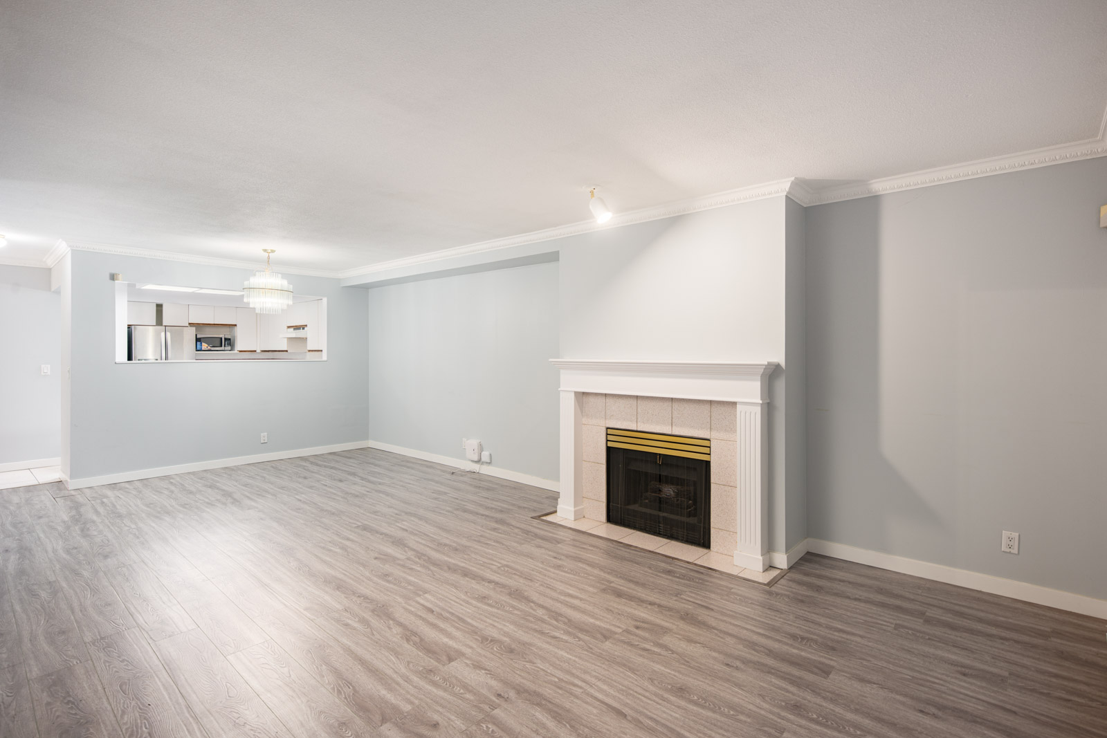 Unfurnished living room with light gray walls, wood flooring, a white fireplace, and an open view into the kitchen area.