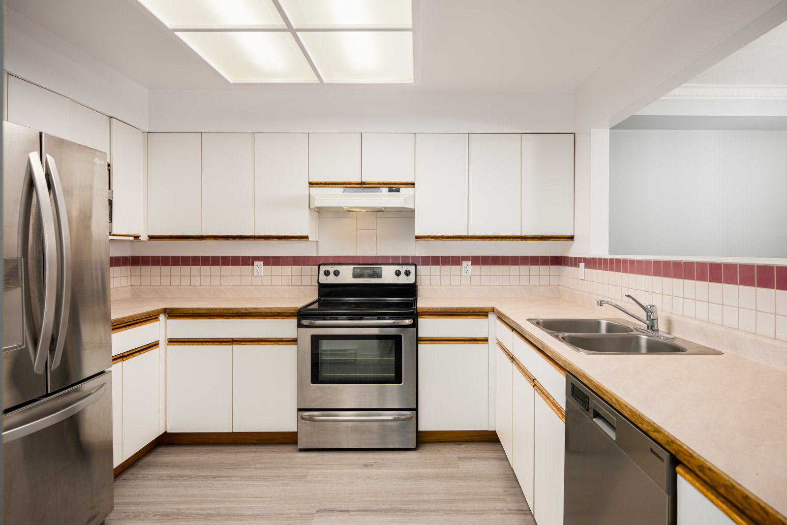 A modern kitchen with white cabinets, stainless steel appliances, a double sink, and beige countertops with pink-tiled backsplash.