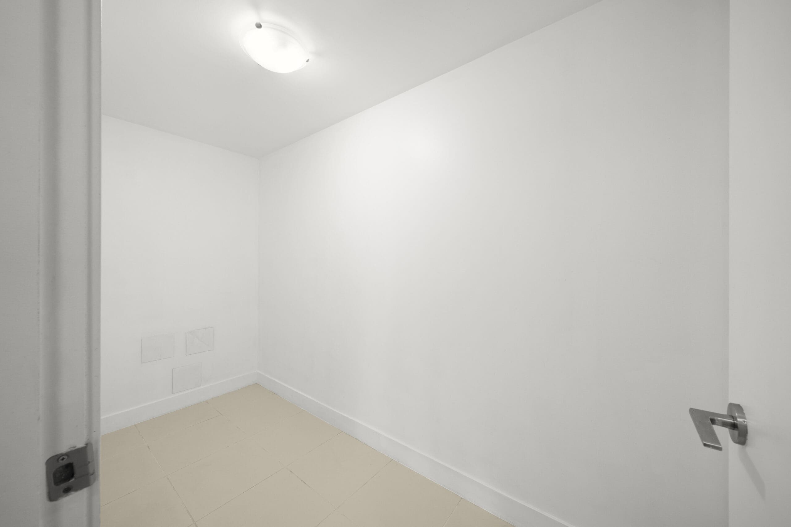 Empty white room with beige tiled floor, plain walls, ceiling light, and partially open door in the foreground.