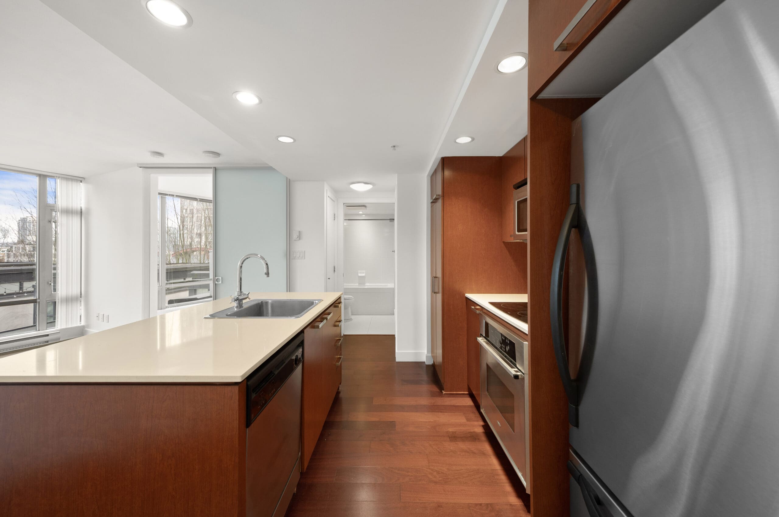 Modern kitchen with stainless steel appliances, wood cabinets, central island with sink, and bright natural light from large windows.