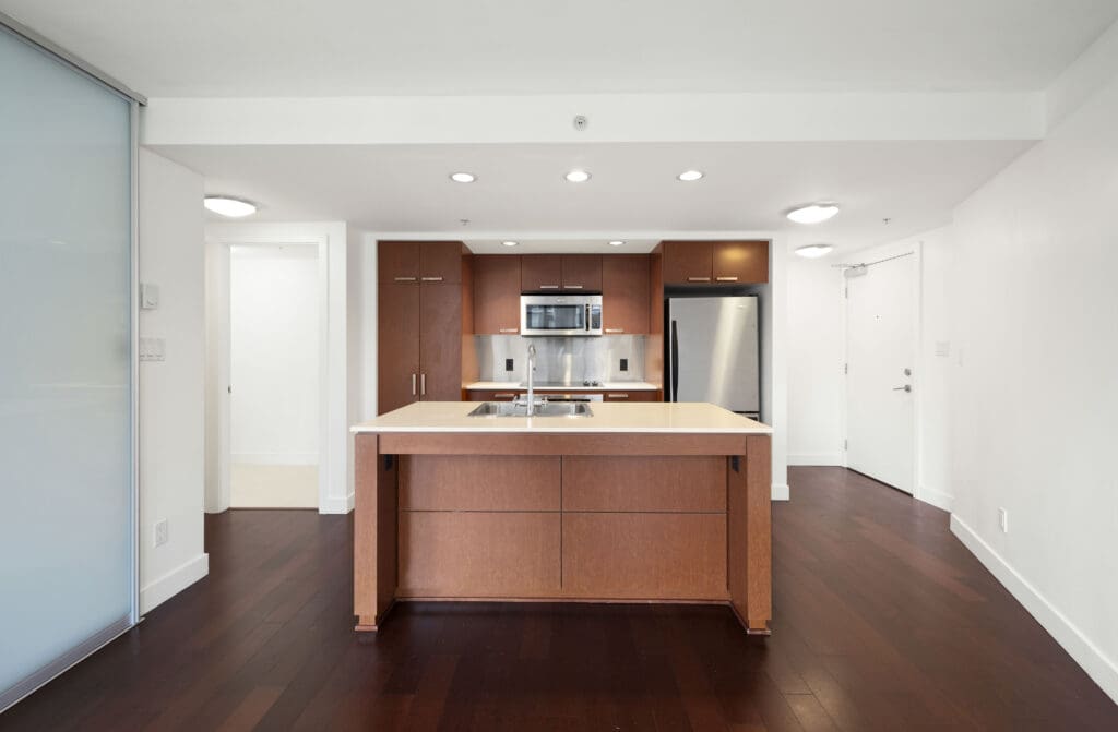 Modern kitchen with brown wooden cabinetry, stainless steel appliances, an island with a double sink, and dark hardwood floors in a bright, open space.