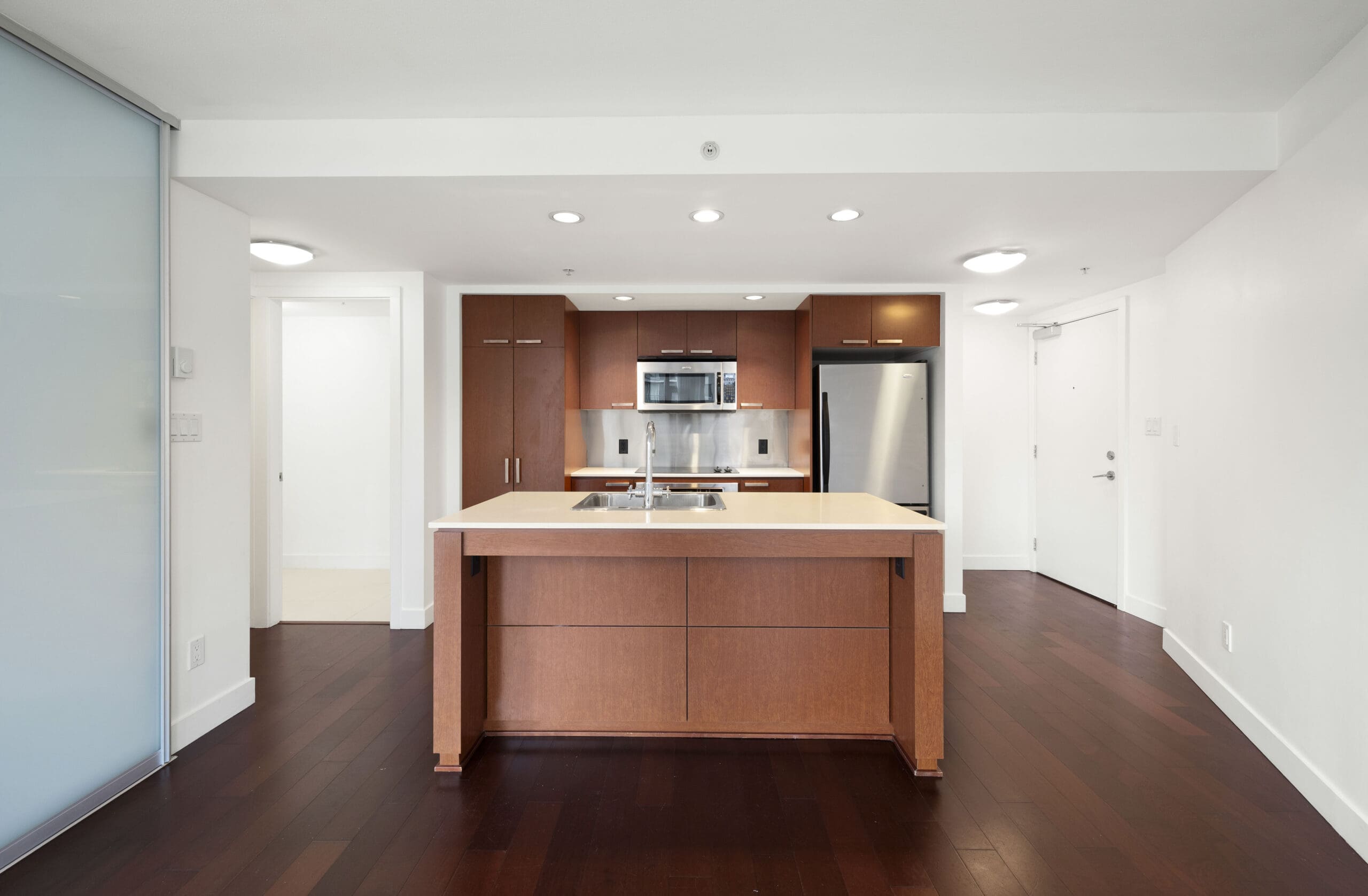 Modern kitchen with brown wooden cabinetry, stainless steel appliances, an island with a double sink, and dark hardwood floors in a bright, open space.