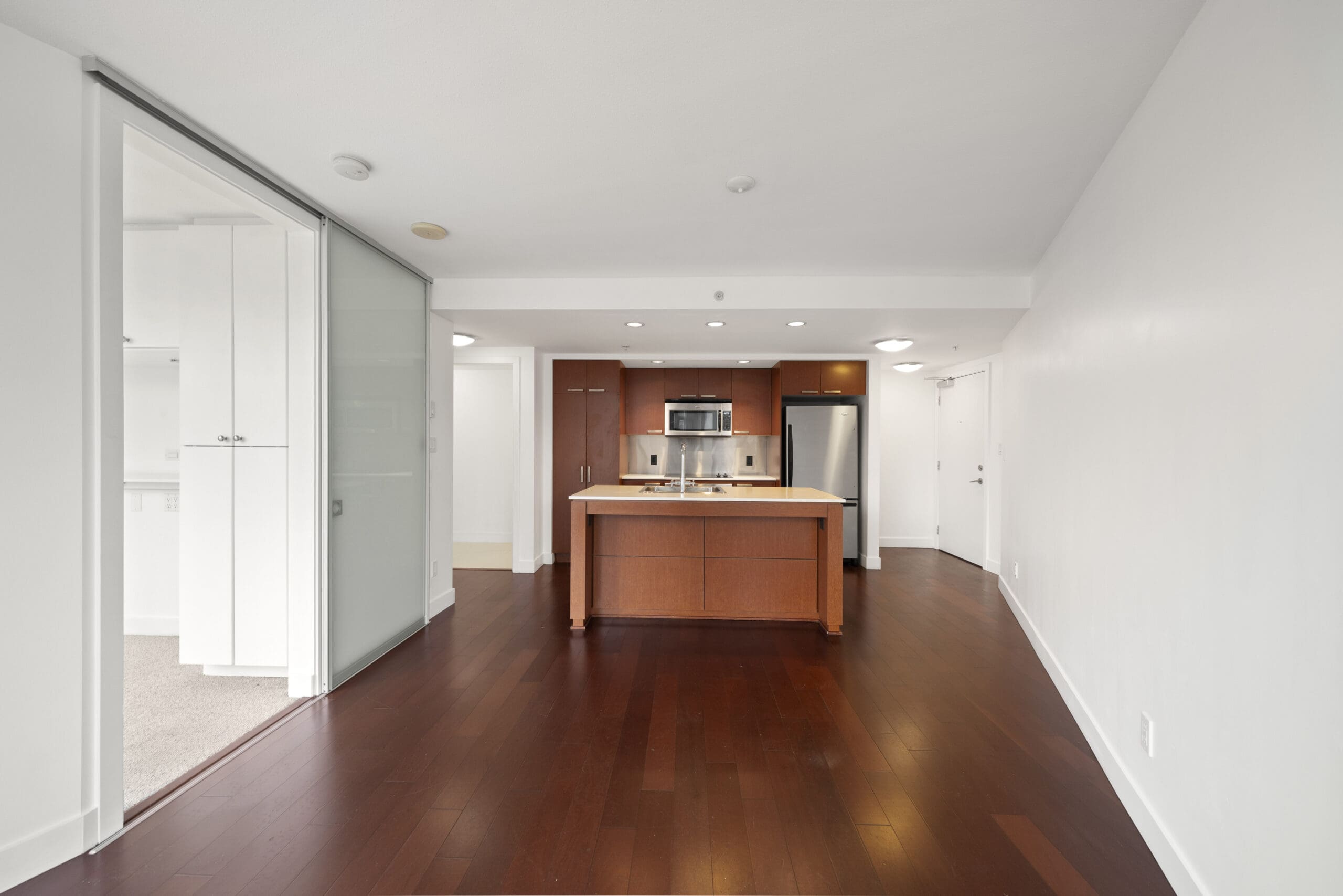 Modern open-plan kitchen with stainless steel appliances, center island, and dark wood floors, adjacent to a glass-walled room and white walls throughout.