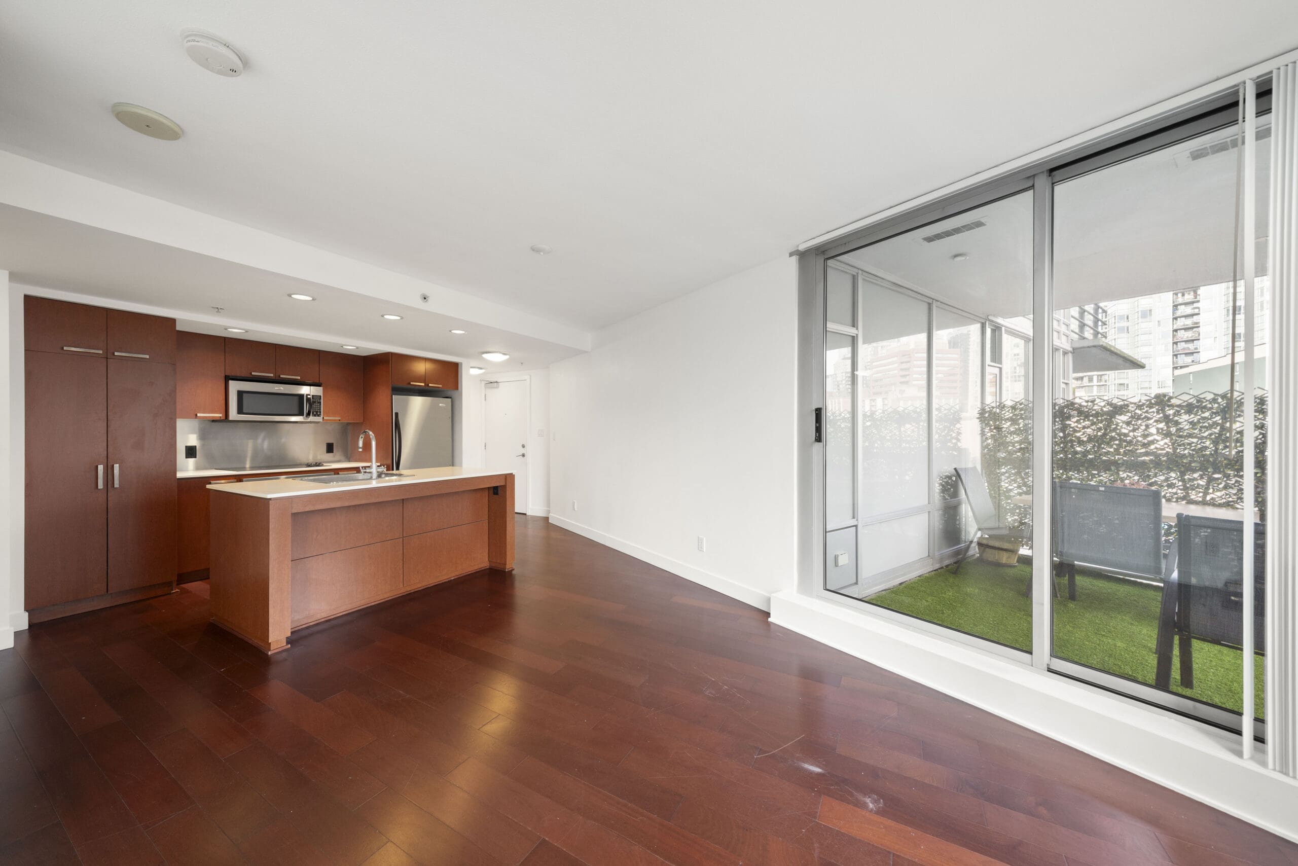 Modern open-plan kitchen and living area with wood flooring, stainless steel appliances, and sliding glass doors leading to a small outdoor patio with artificial grass and patio furniture.