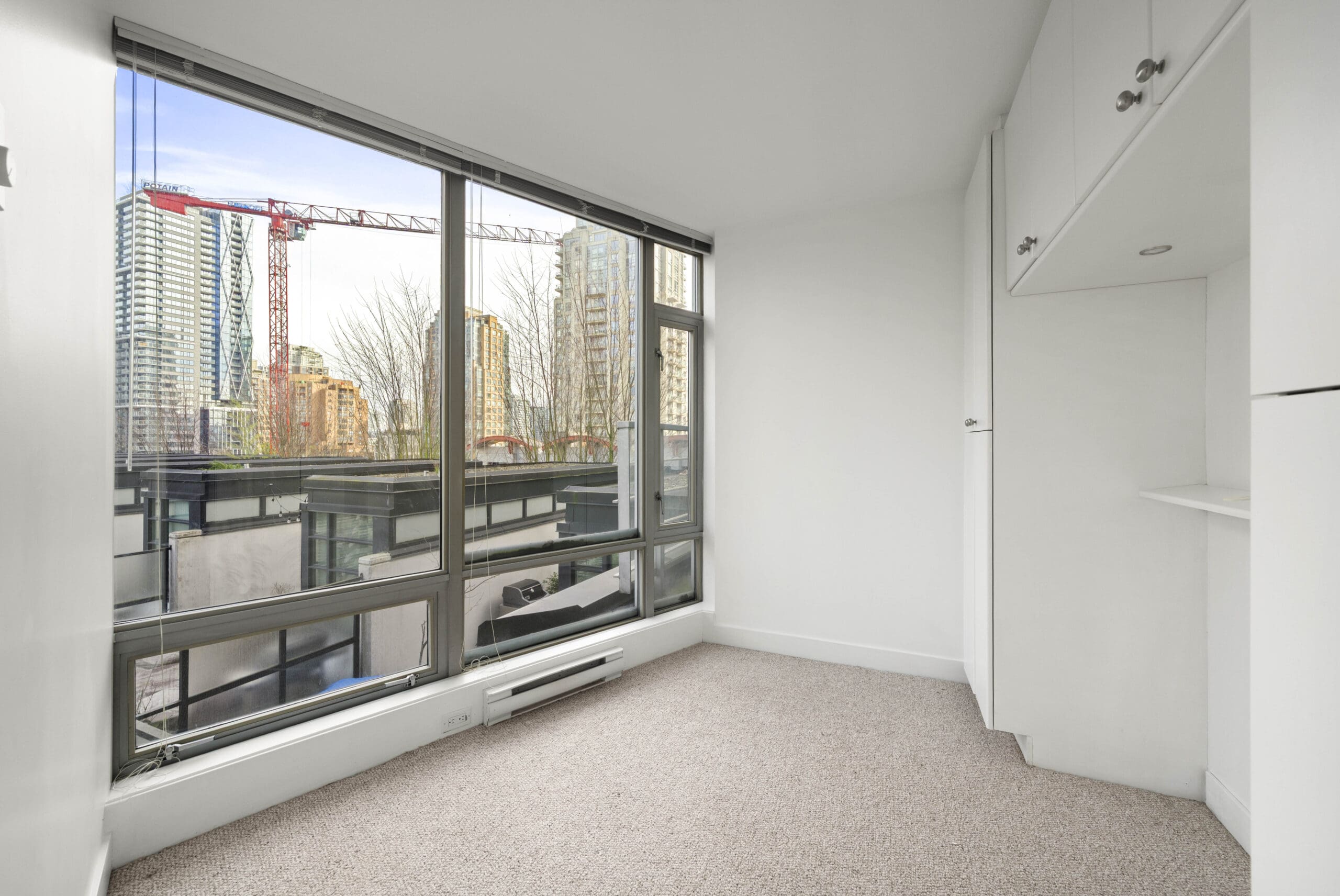 Small, unfurnished room with large windows overlooking city buildings and a construction crane; white walls, built-in shelving, and beige carpeted floor.