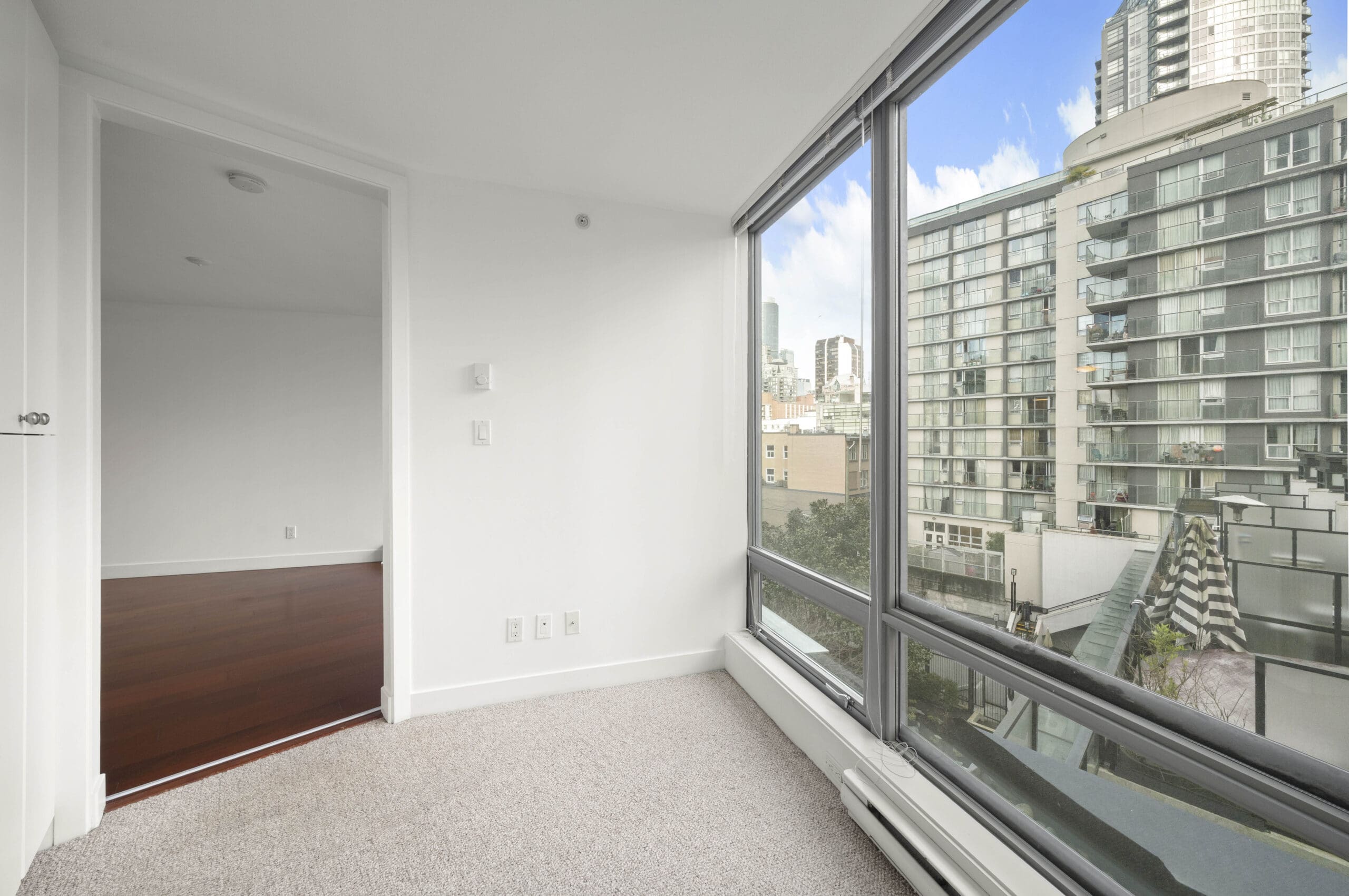 Corner of a modern apartment with white walls, carpeted floor, and large windows overlooking neighboring high-rise buildings.