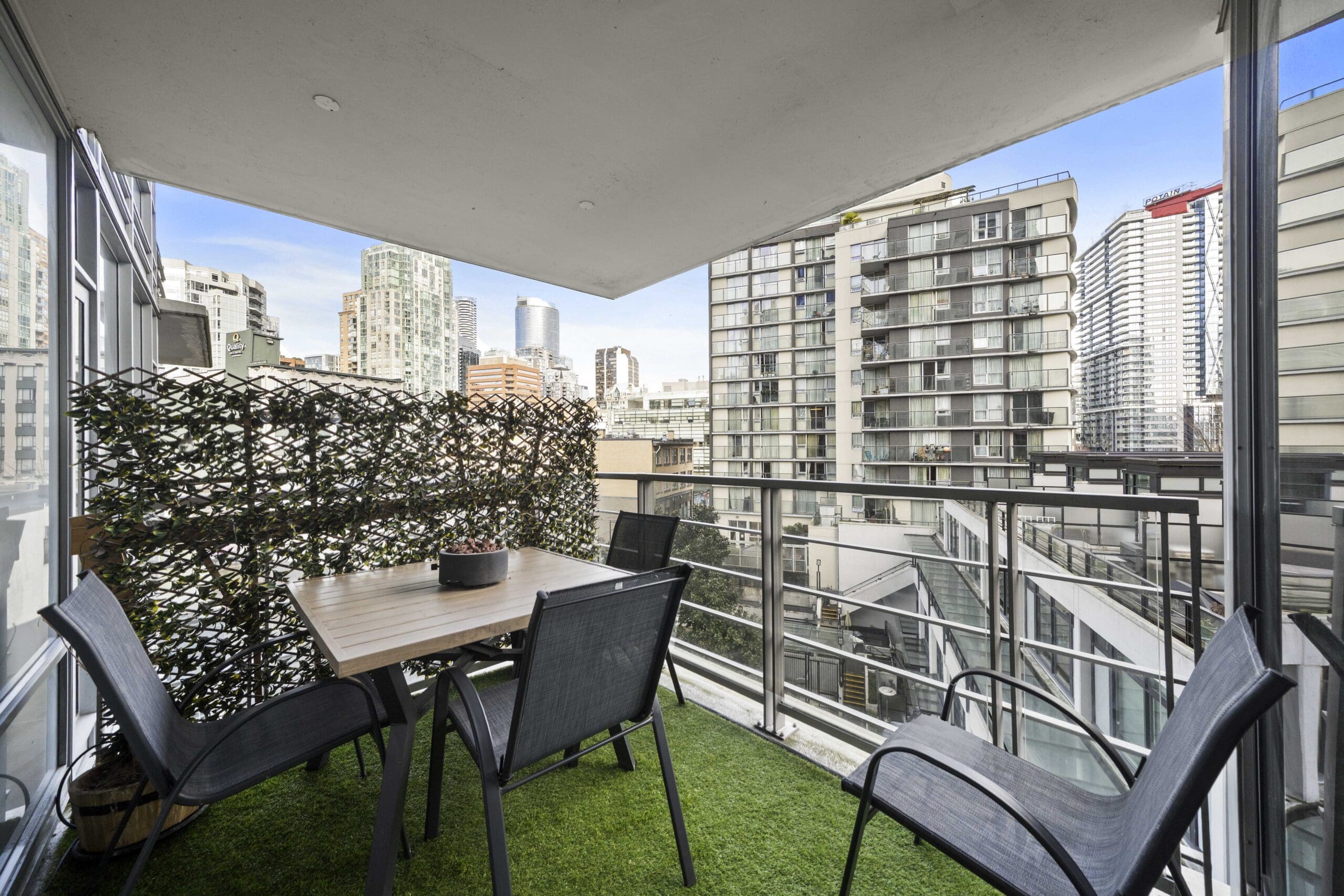 Modern balcony with artificial grass, a table and four chairs, surrounded by glass railings, overlooking city high-rise buildings.