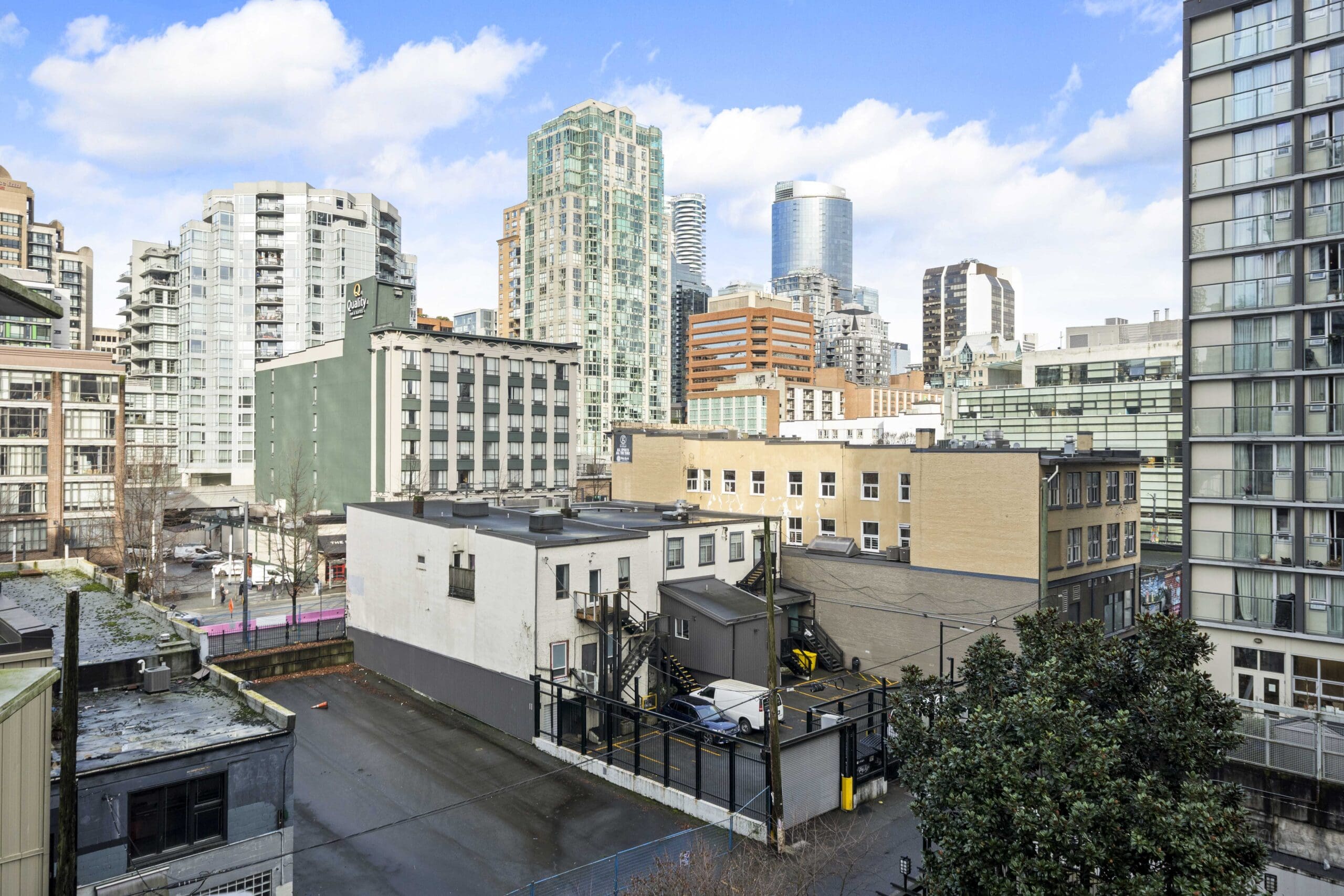 View of a cityscape featuring mid-rise and high-rise buildings, with a parking area, alley, and some greenery in the foreground under a partly cloudy sky.
