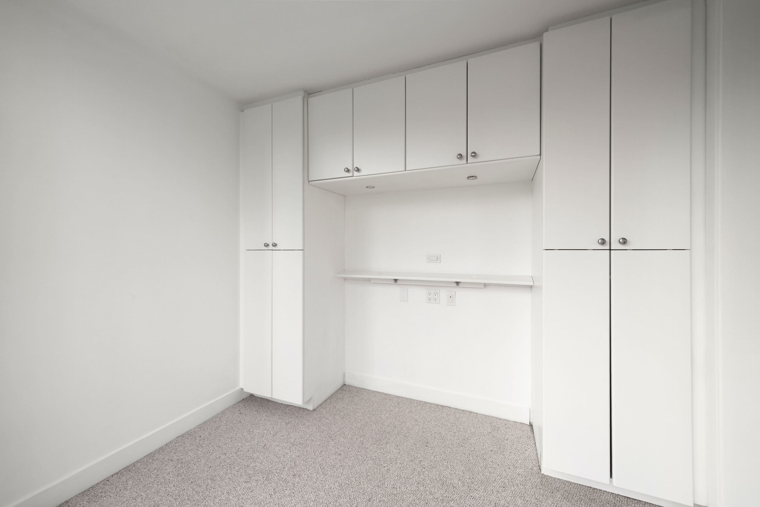 Minimalist room with white built-in cabinets, an empty alcove, beige carpet, and white walls. No furniture or decorations are present.