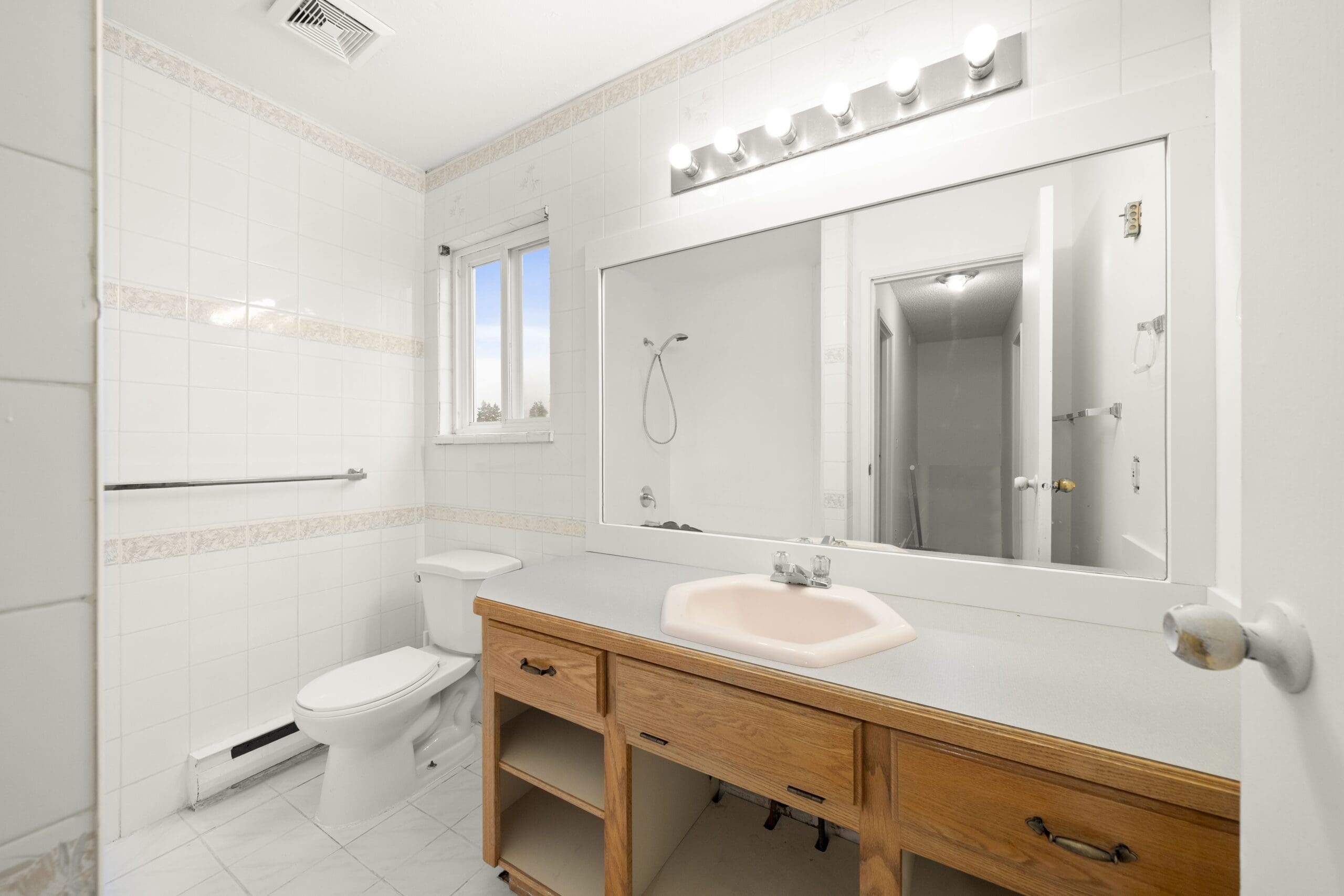 Bathroom with large wall mirror, white tile walls, a window, toilet, light wood vanity with pink sink, and exposed plumbing beneath the countertop.