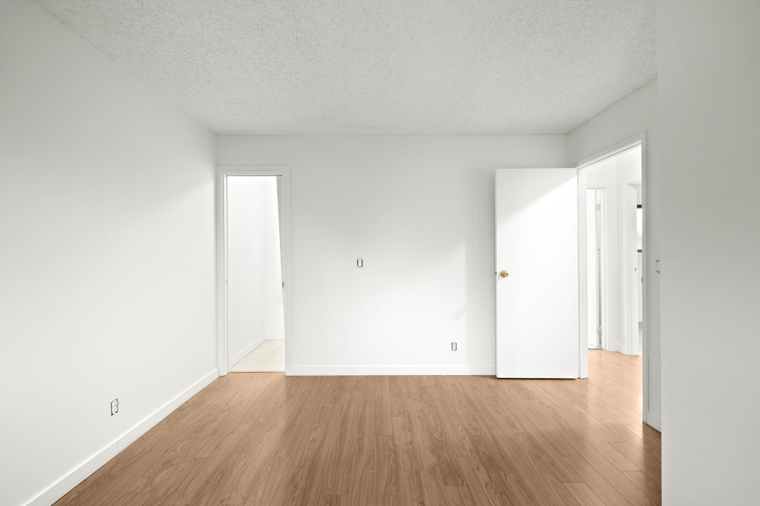 Empty room with white walls, light wood flooring, an open door, a closed door, and a doorway leading to a tiled area.
