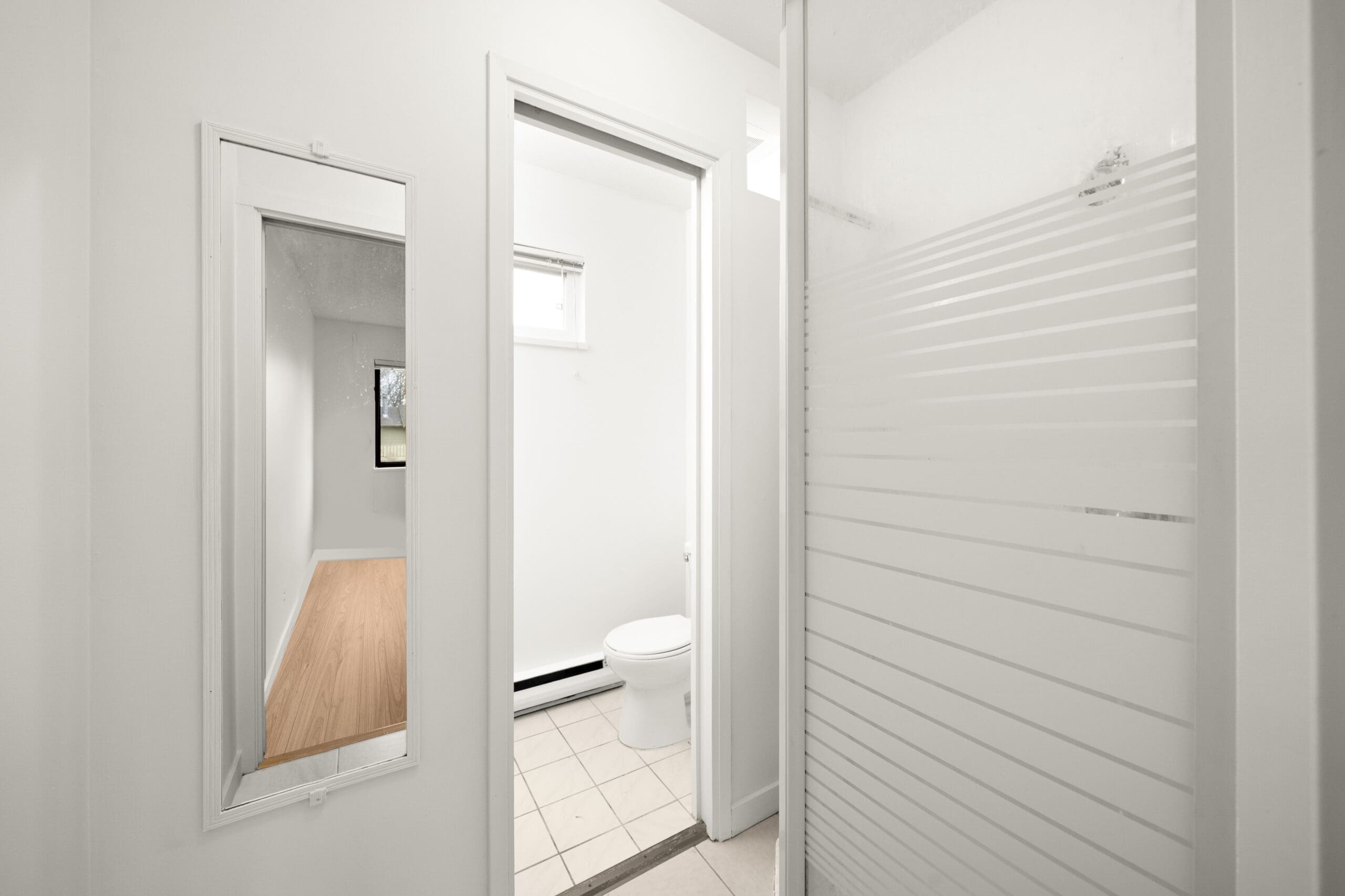 A small bathroom with white walls, a toilet, a shower with a frosted glass door, a tile floor, and an open door leading to a room with wood flooring and a window.