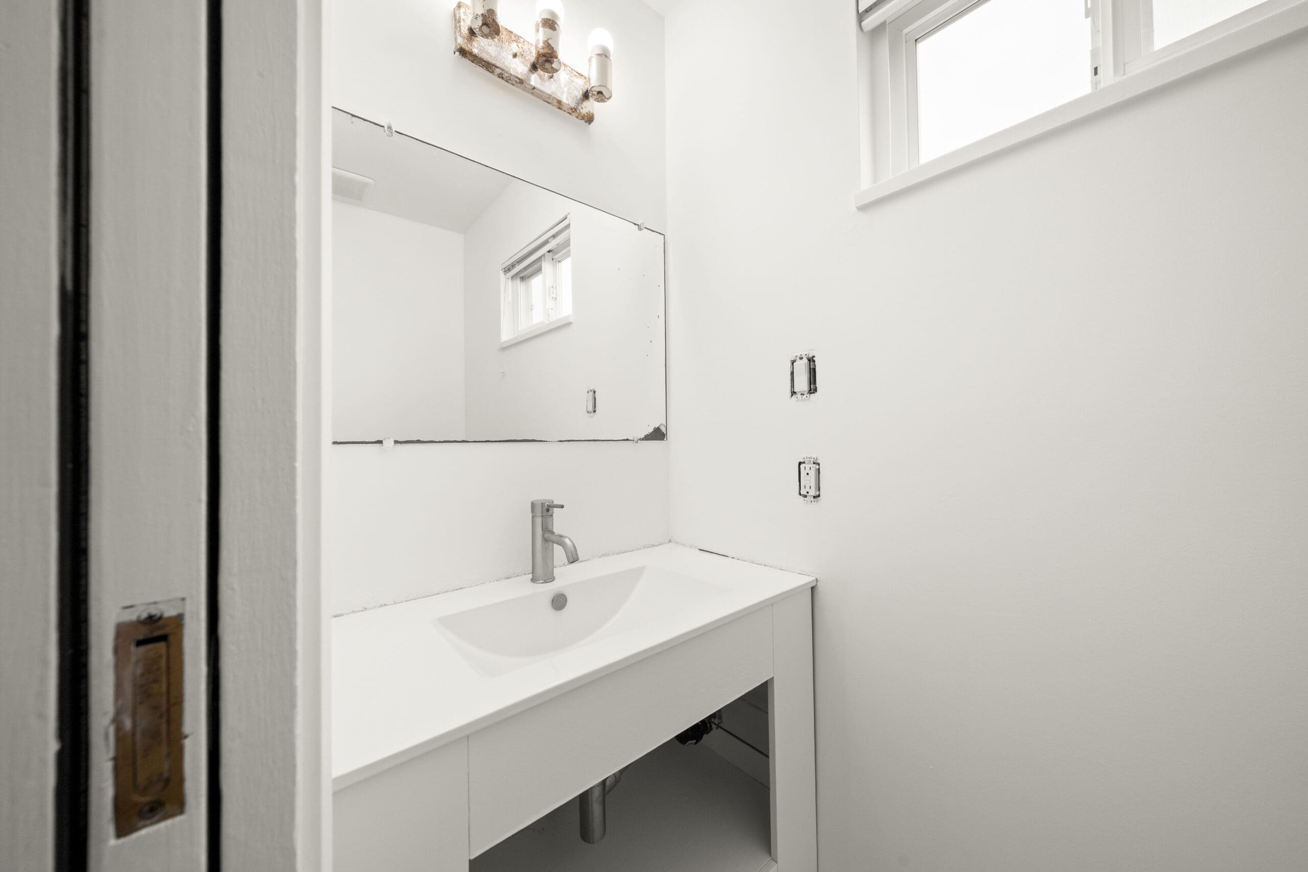 A white bathroom with a rectangular mirror, modern sink, exposed plumbing, light fixture above, and two windows. Electrical outlets appear unfinished.