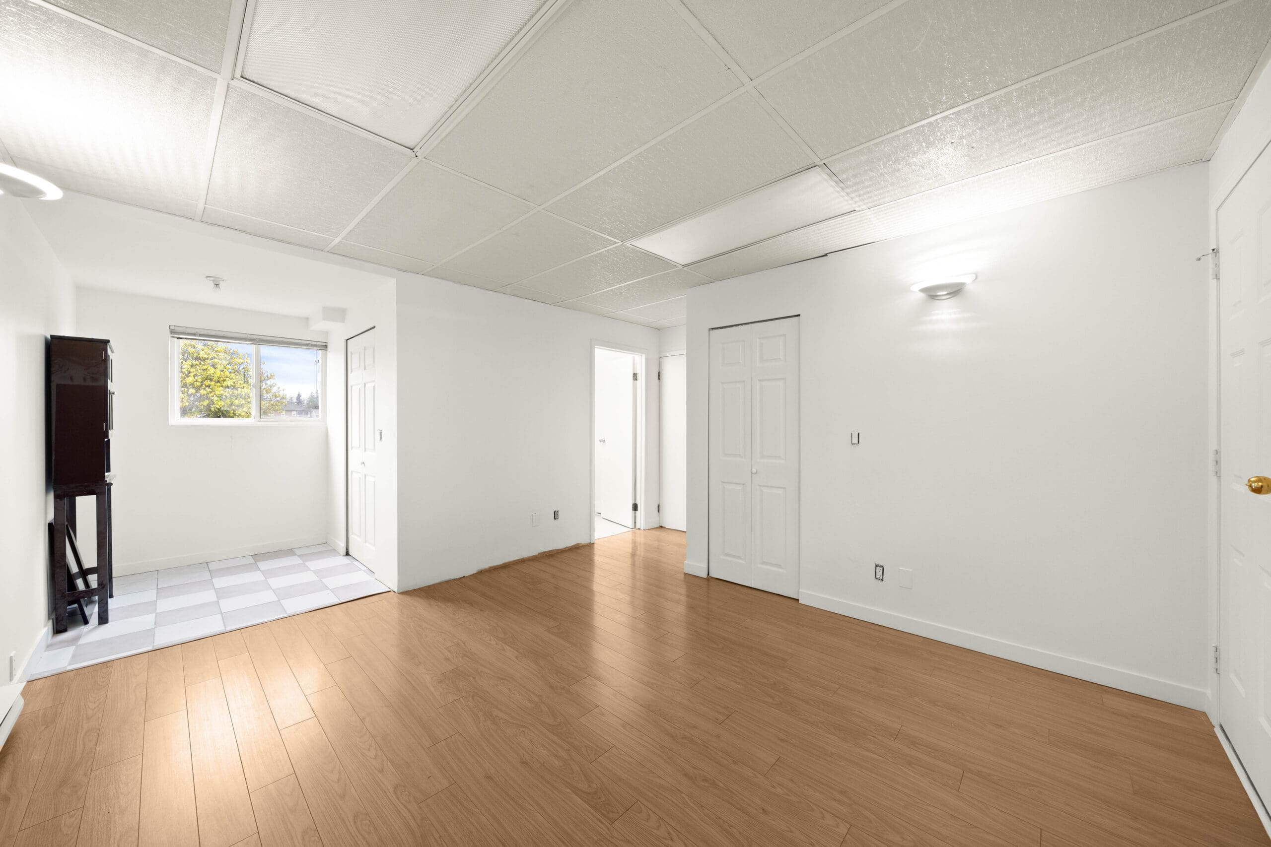 Bright, empty room with white walls, wood flooring, and a white door; adjoining a smaller tiled area with a window.