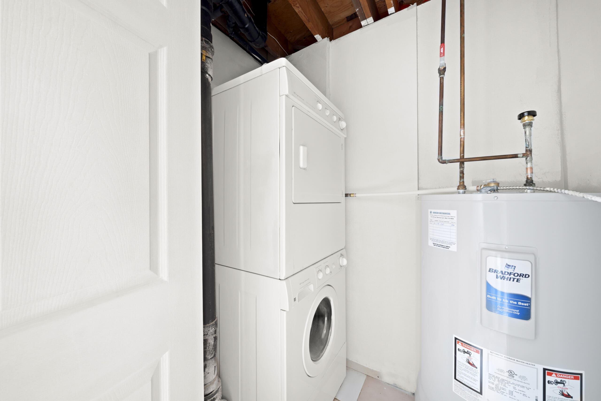 A stacked washer and dryer unit is positioned next to a water heater in a small utility room with exposed ceiling beams.