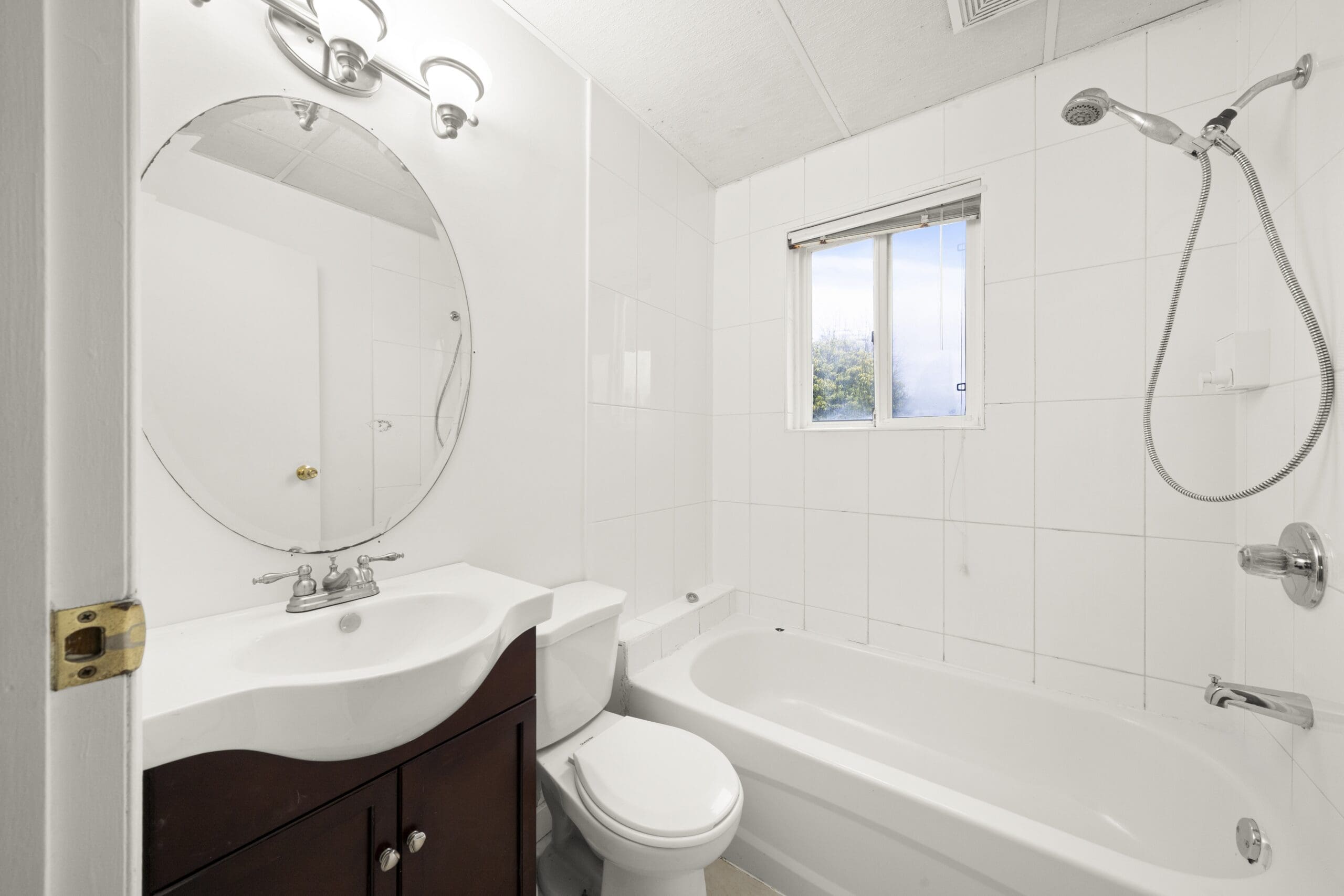 A white-tiled bathroom with a vanity and sink, oval mirror, toilet, bathtub with showerhead, and a window letting in natural light.