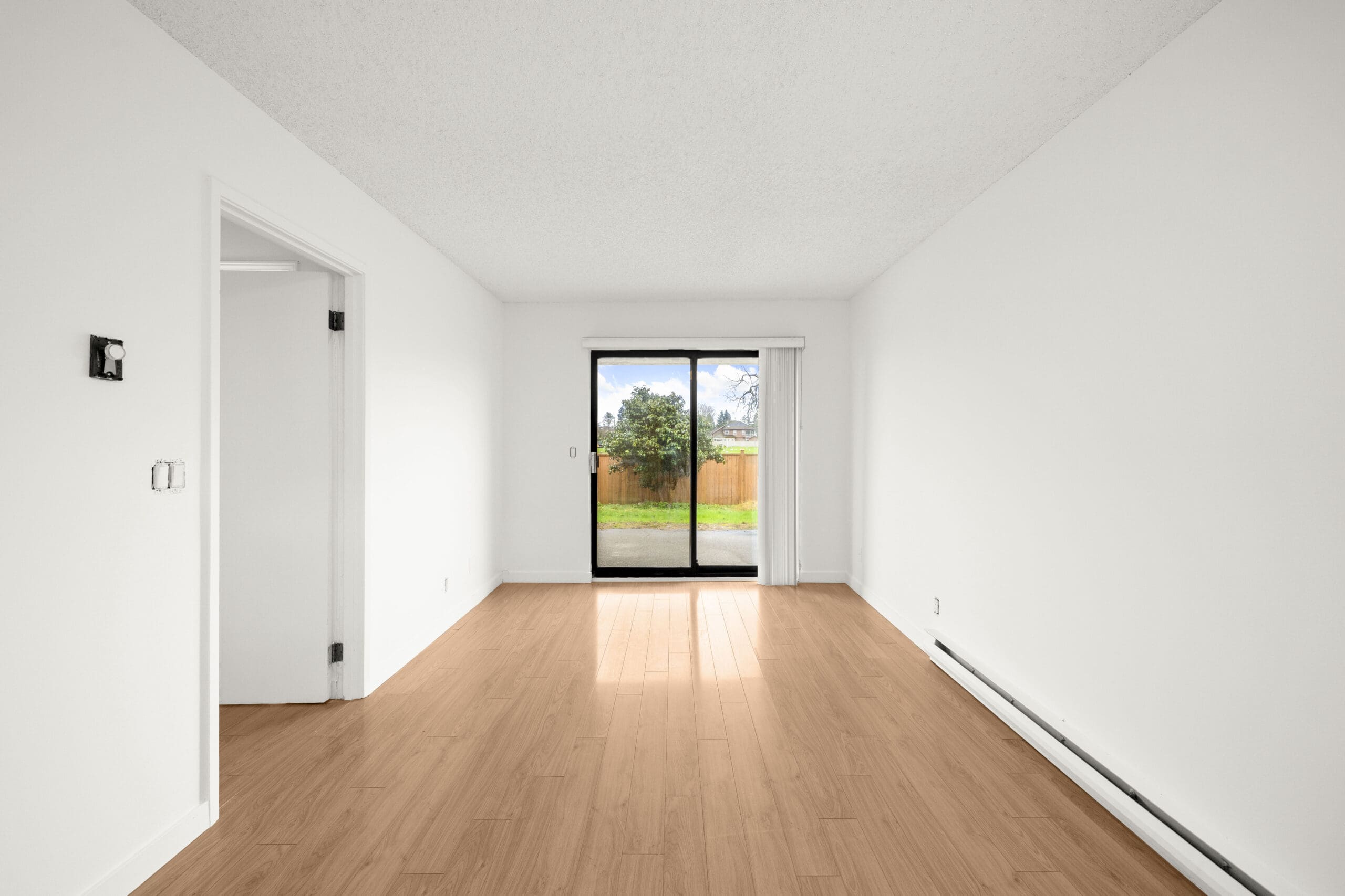 Empty, unfurnished room with white walls, light wood flooring, a sliding glass door leading to a fenced yard, and an open doorway to the left.
