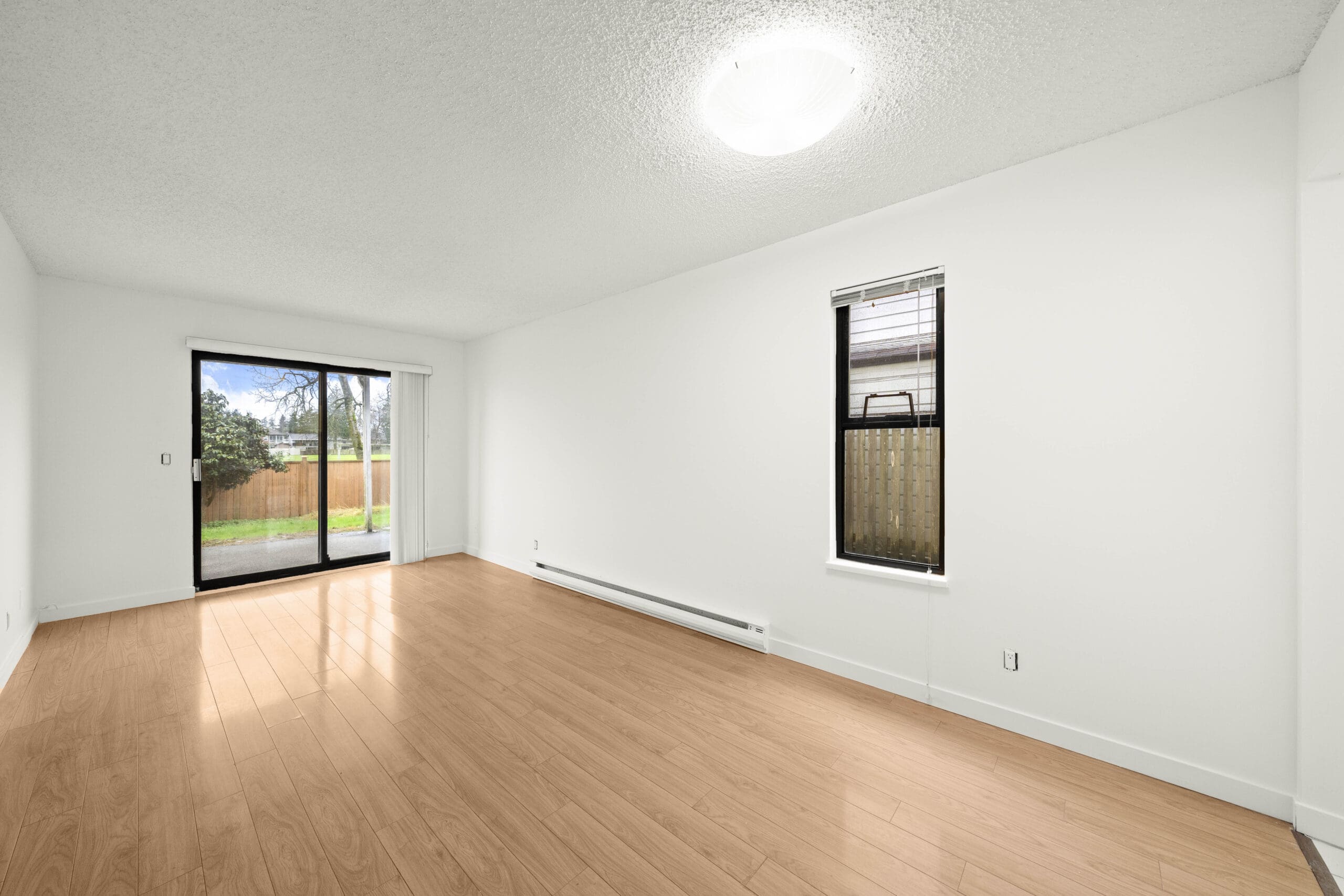 Empty room with light wood flooring, white walls, a window with blinds, and a sliding glass door leading to a fenced yard.
