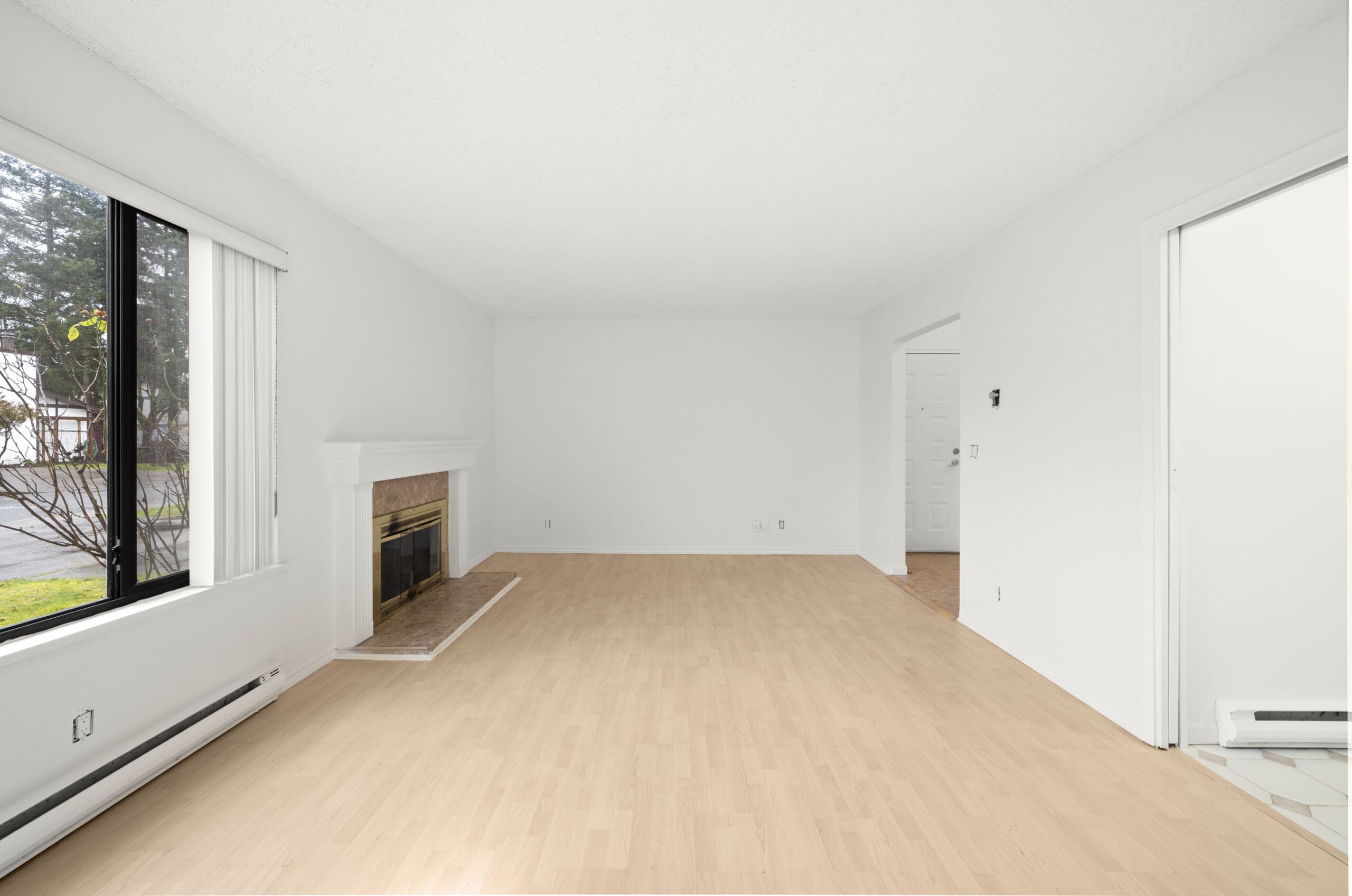 Unfurnished living room with light wood flooring, white walls, a fireplace on the left wall, large window, and open doorways leading to other rooms.