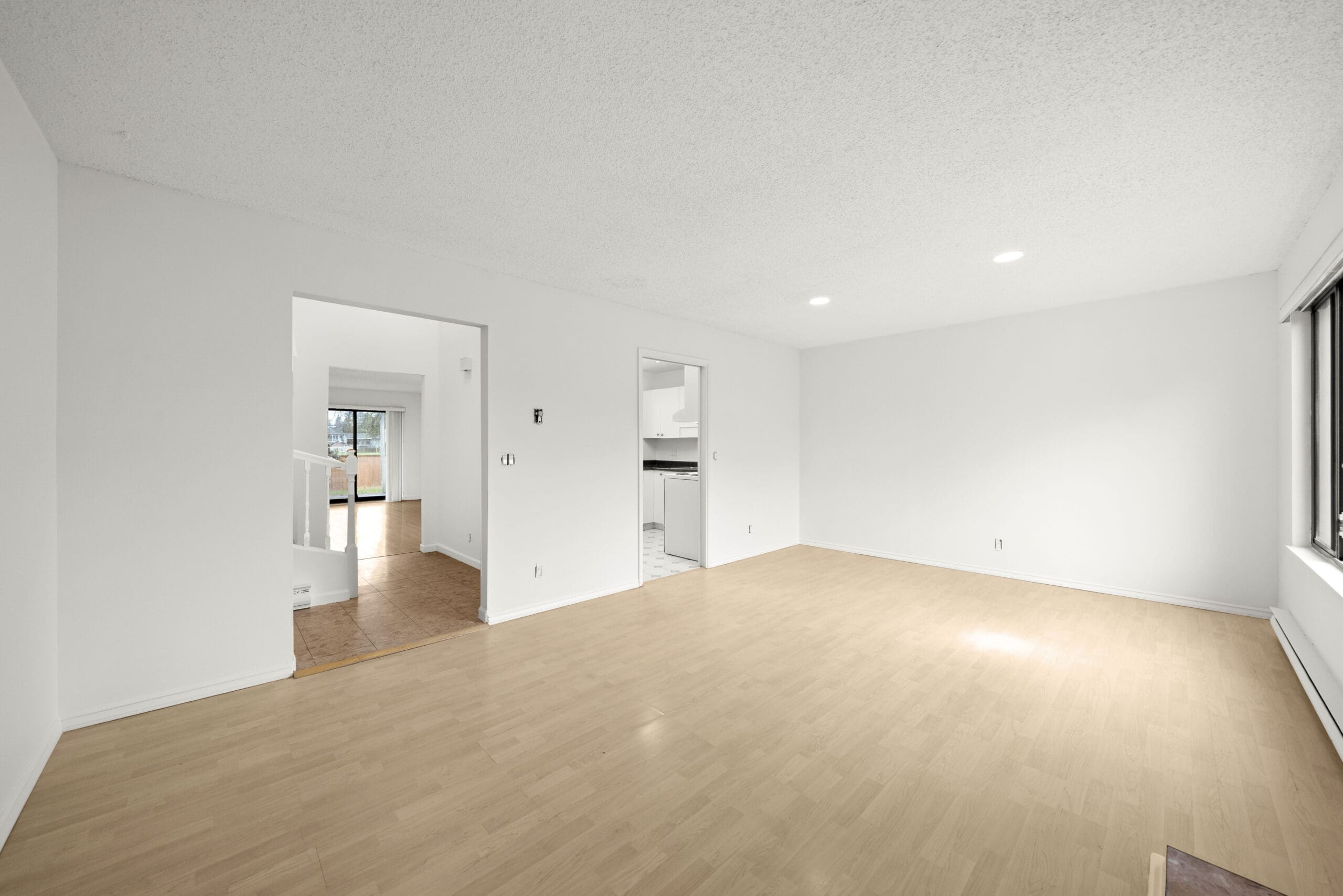 Empty room with light wood flooring, white walls, recessed ceiling lights, large window on the right, and open doorways leading to adjacent rooms.
